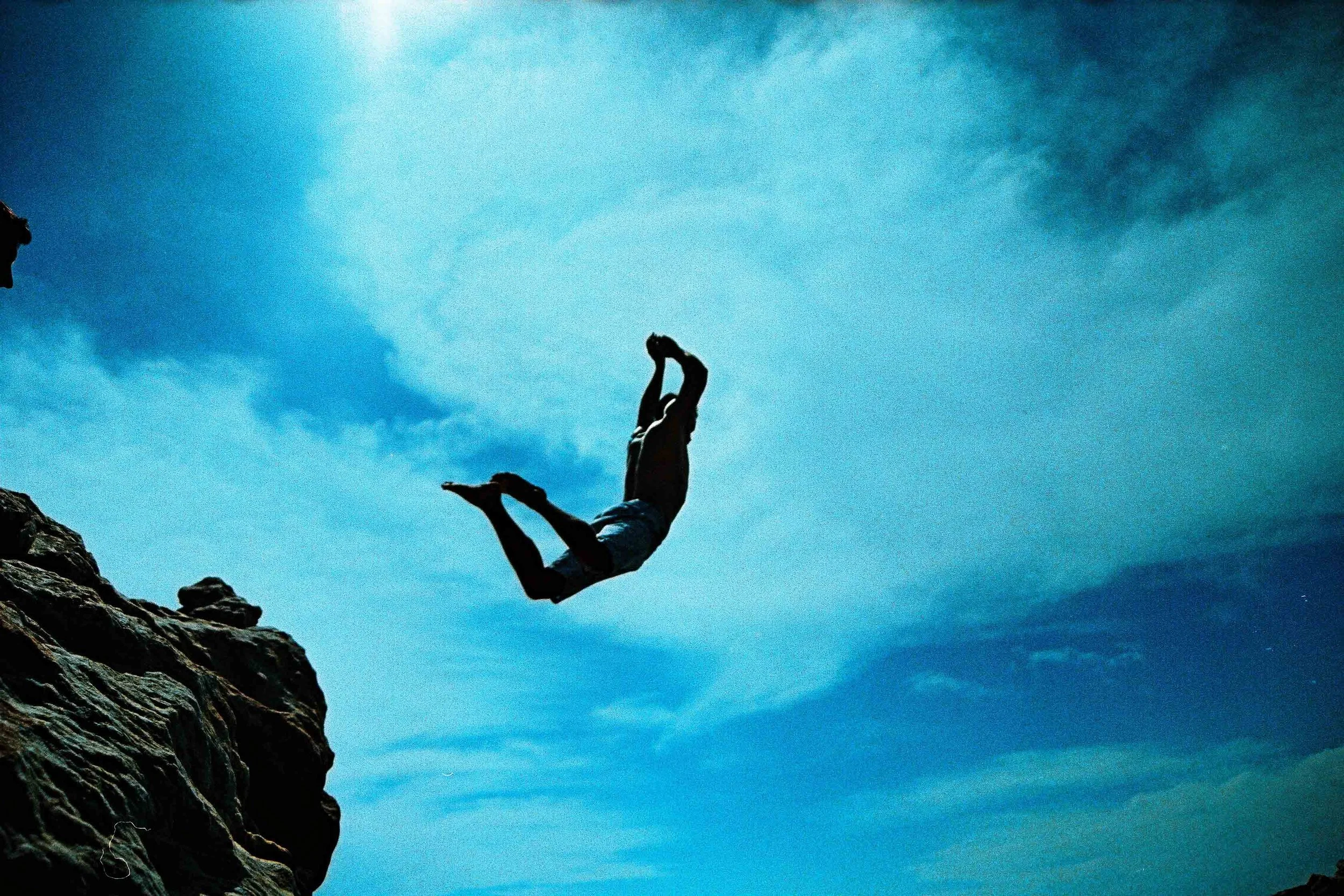 A person falling backwards off a rocky cliff into the air with a blue sky and clouds in the background.