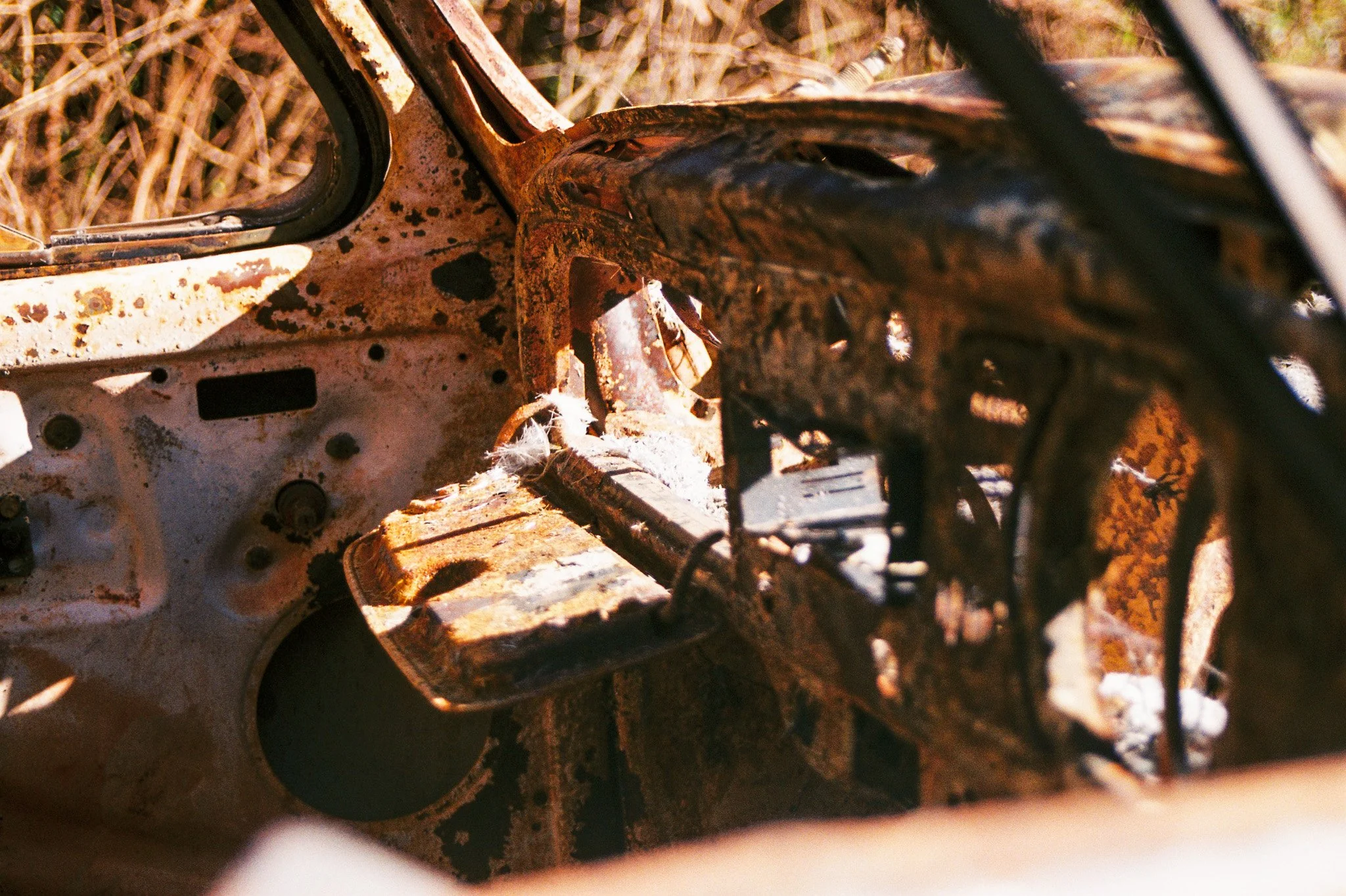 Close-up of a rusty, weathered, and partially disassembled vintage car or vehicle part, surrounded by dry grass.