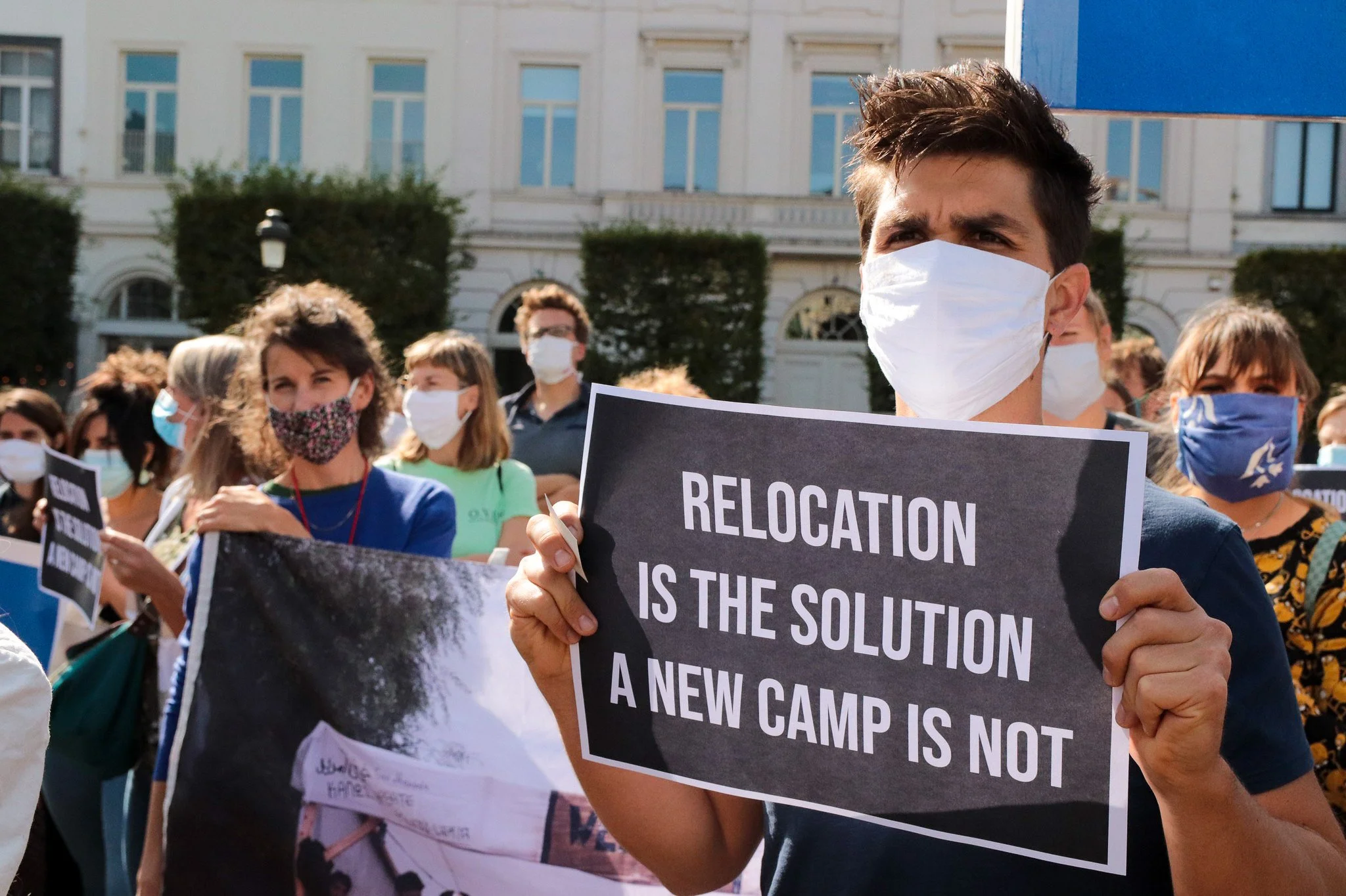 Protesters wearing face masks, holding signs, with a building with windows and trees in the background.