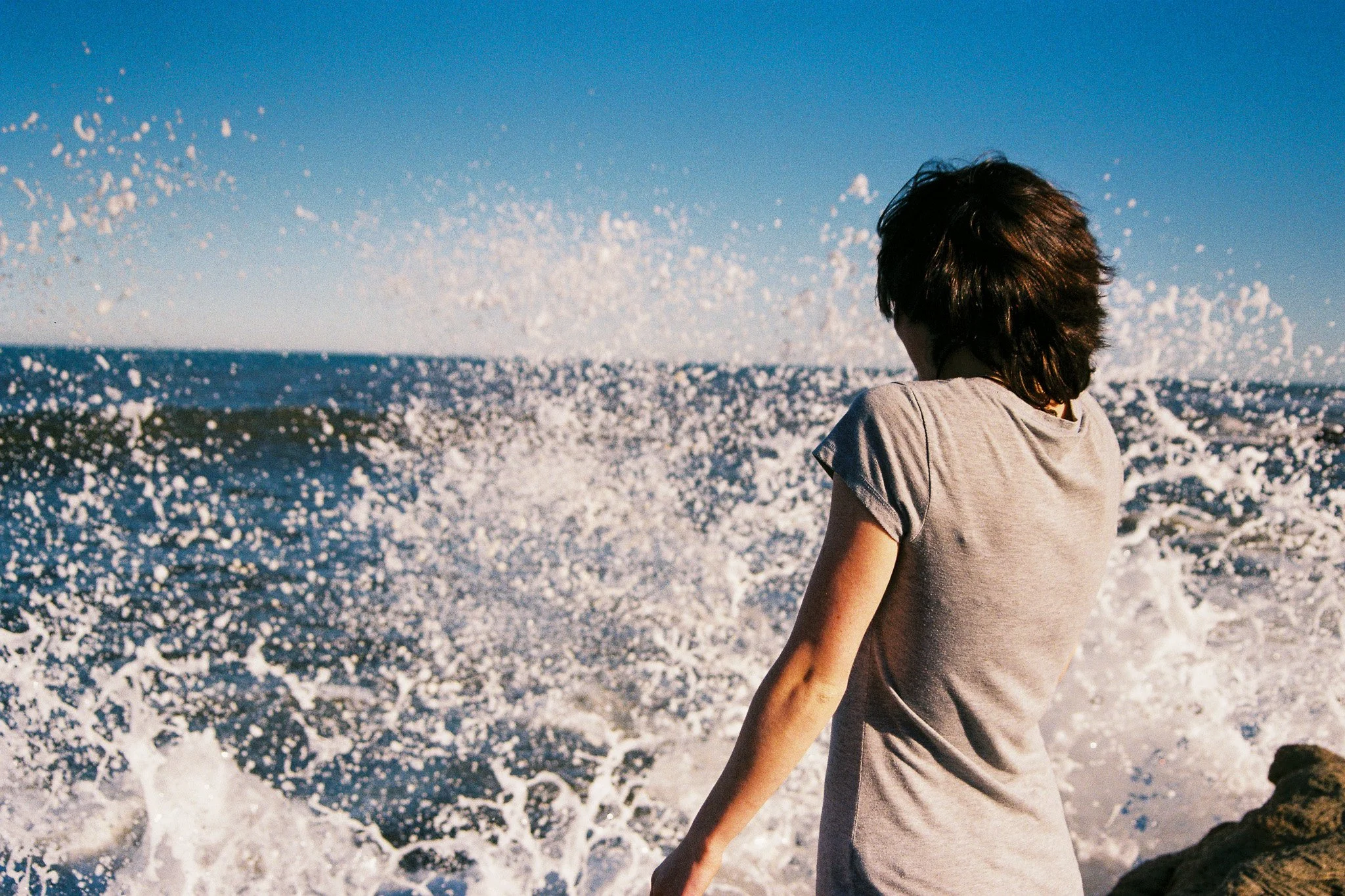 Person with shoulder-length hair wearing a gray T-shirt standing near the ocean, facing away, with waves crashing behind them.