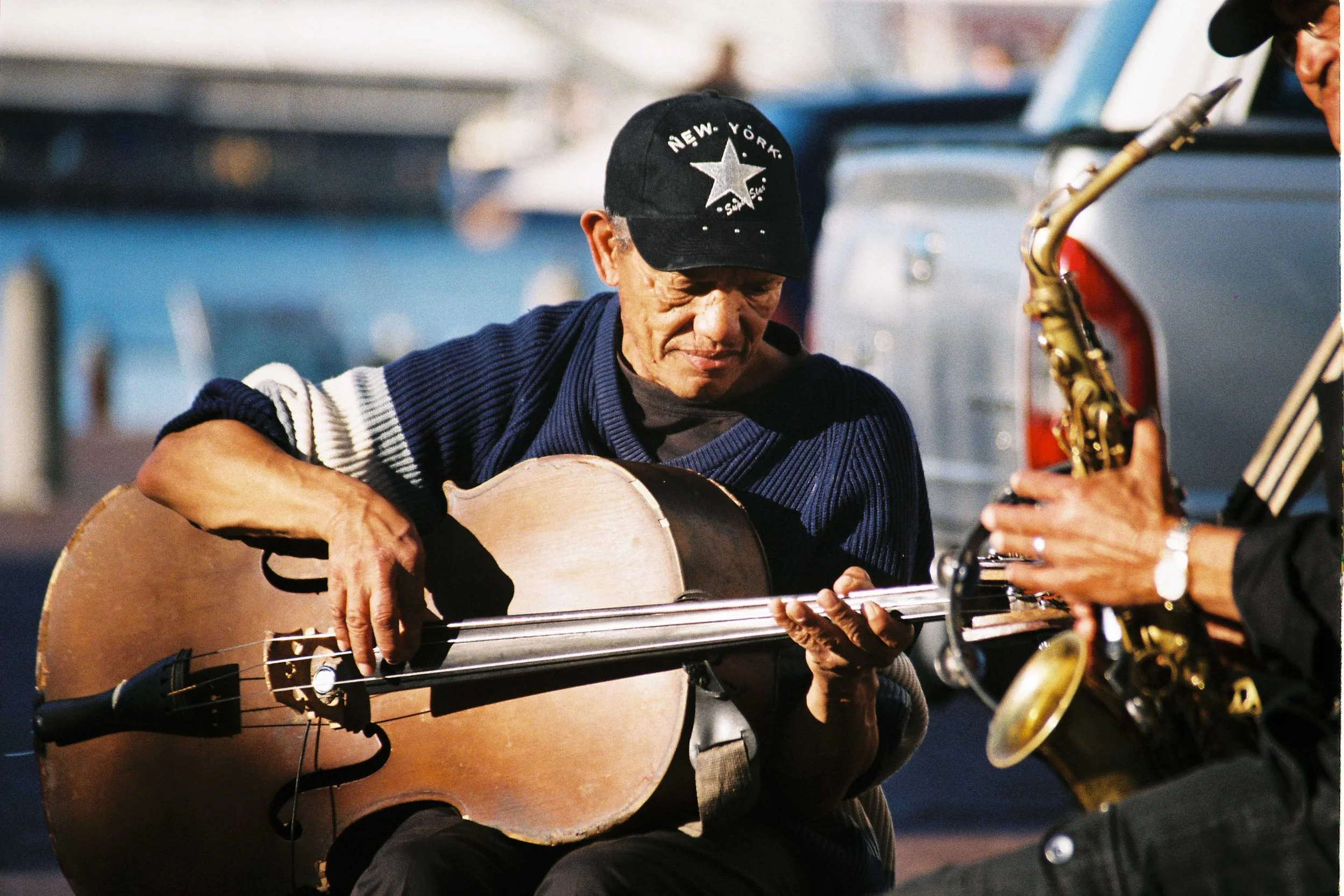 A man wearing a black cap with a star logo is playing an cello like an acoustic guitar in an outdoor setting. Another musician is partially visible playing a saxophone nearby.