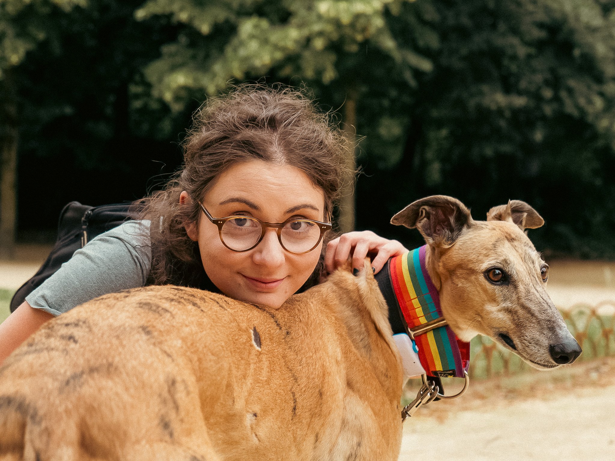 A woman with glasses and curly hair in a park resting her head on a large dog with tan fur and a rainbow striped collar.