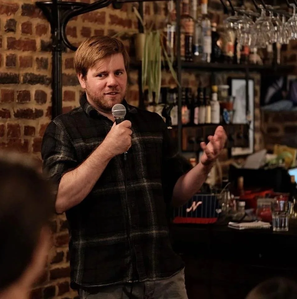 A man with light brown hair and beard, wearing a black plaid shirt, holding a microphone and speaking on stage in a dimly lit bar or restaurant with brick walls and shelves of bottles and glasses.