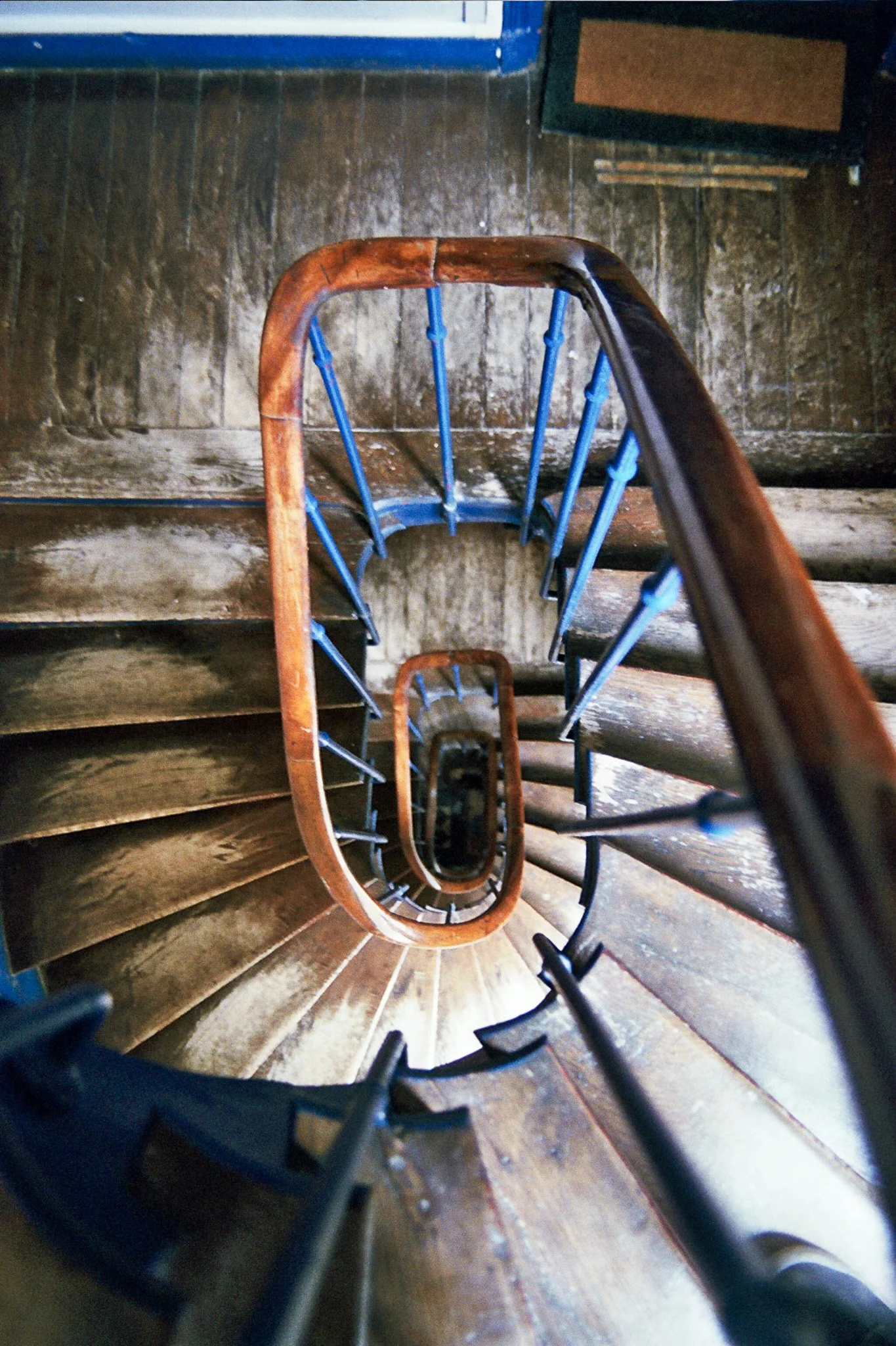 Looking down a spiral staircase with wooden steps and a decorative black metal railing, with a worn wooden wall visible at the top.