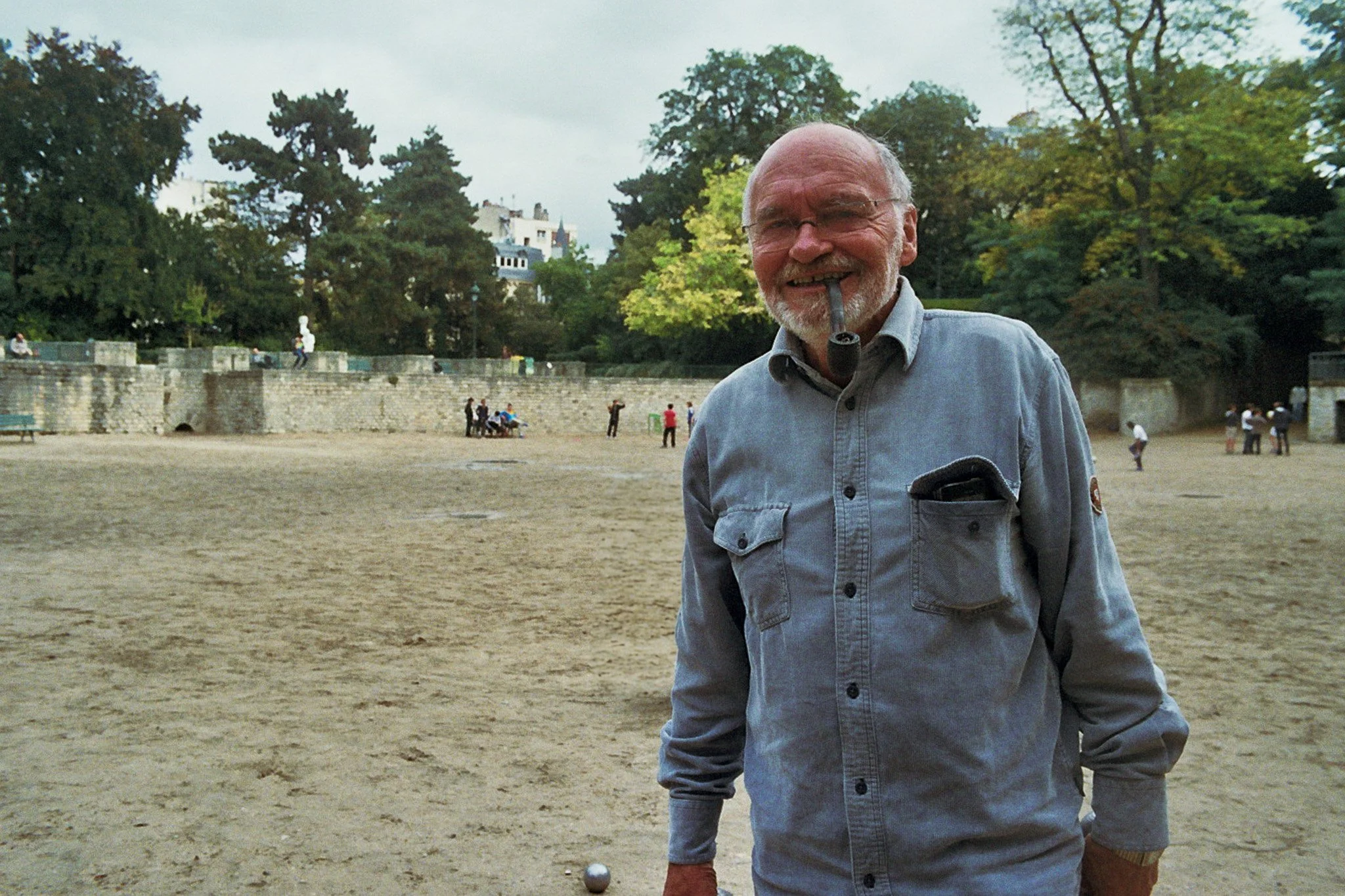 An elderly man with a beard and glasses, wearing a light gray button-up shirt, stands outdoors near a lawn bowl game, with trees and historic buildings in the background.