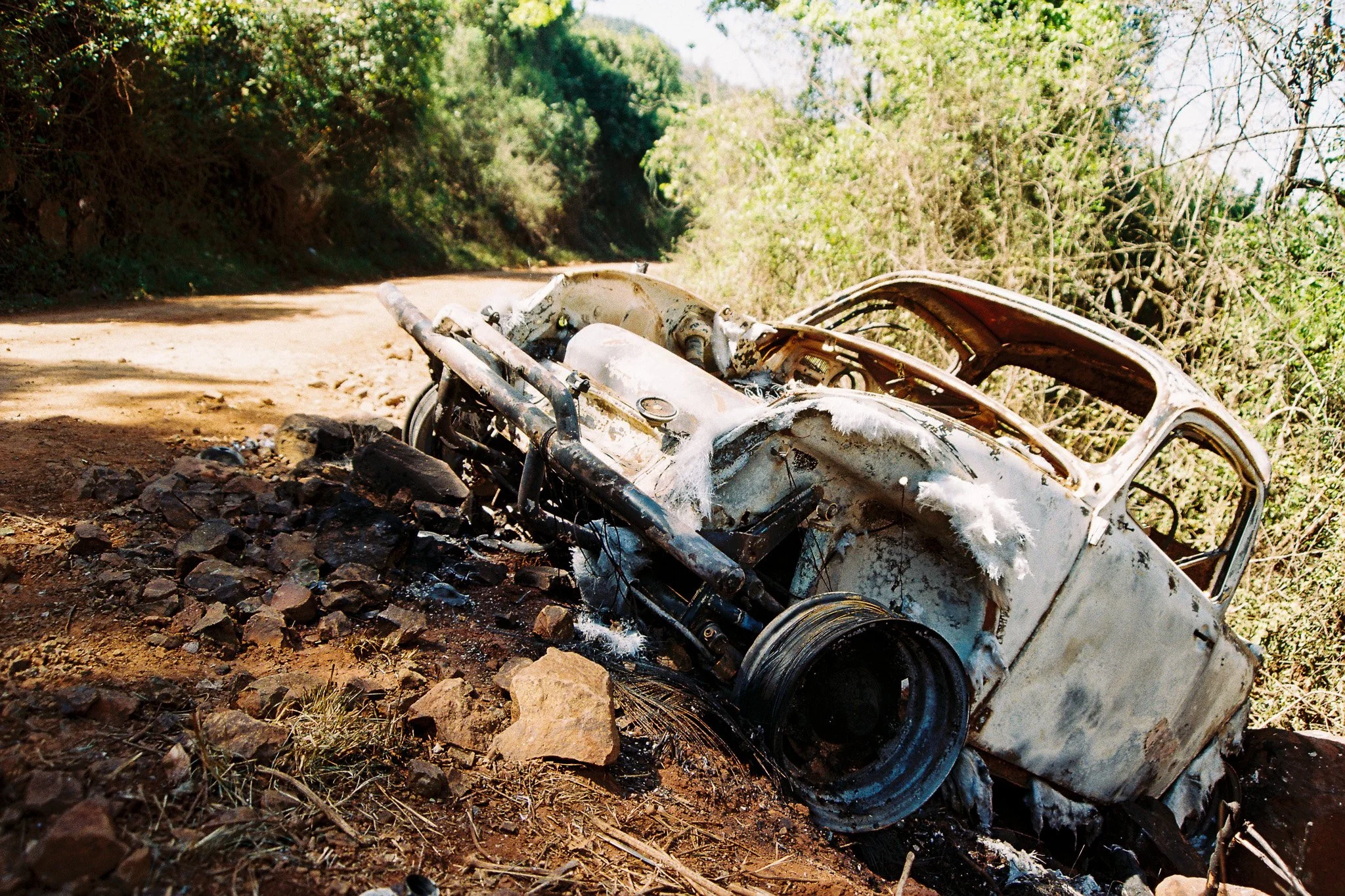 A destroyed white vehicle lying burned and rusted on a dirt road, surrounded by rocks and green bushes, with trees in the background.
