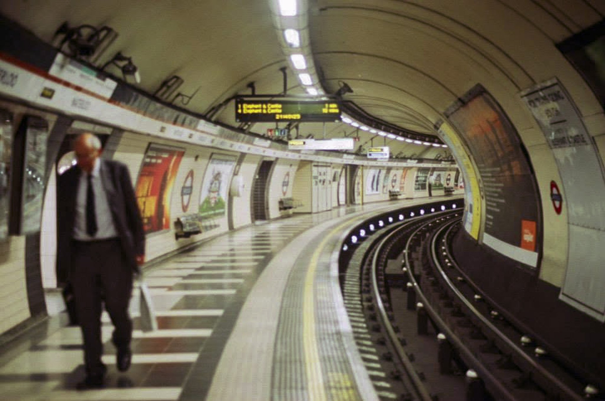 An underground subway station with a man in a suit walking along the platform. The station has a curved ceiling, illuminated signs, and advertisements on the walls.