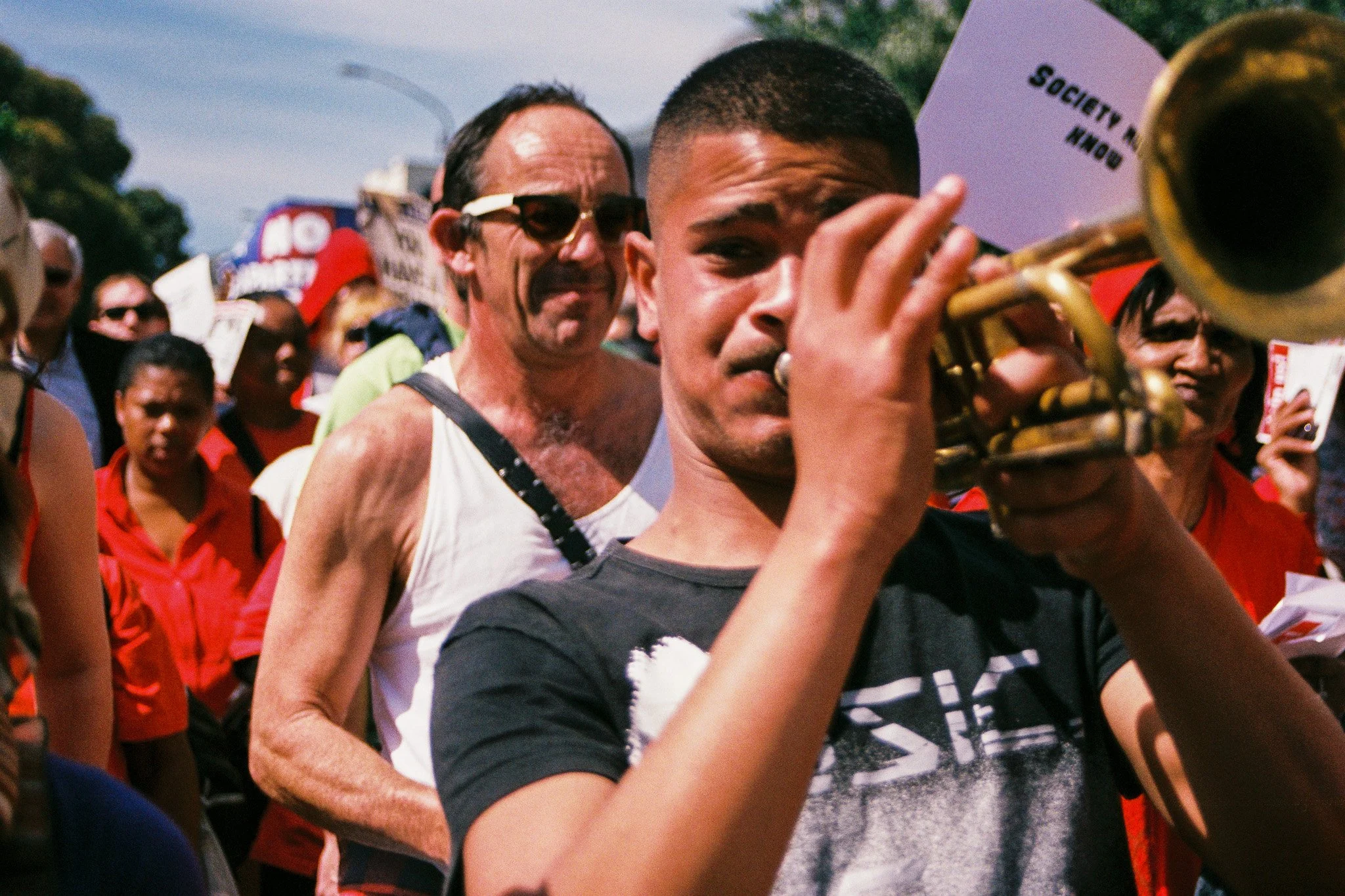 A group of diverse people participating in a protest or rally, with a man in the foreground holding a trumpet and a sign that reads 'Society' in the background.