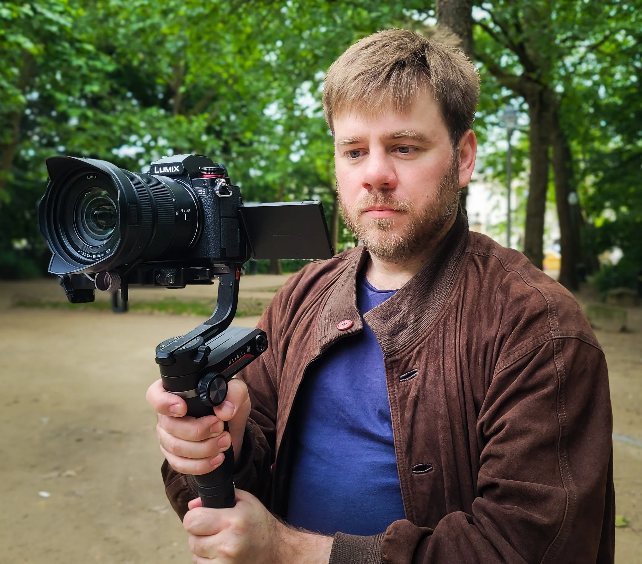 A man with short blond hair and a beard holds a camera mounted on a stabilizer in an outdoor park with green trees in the background.