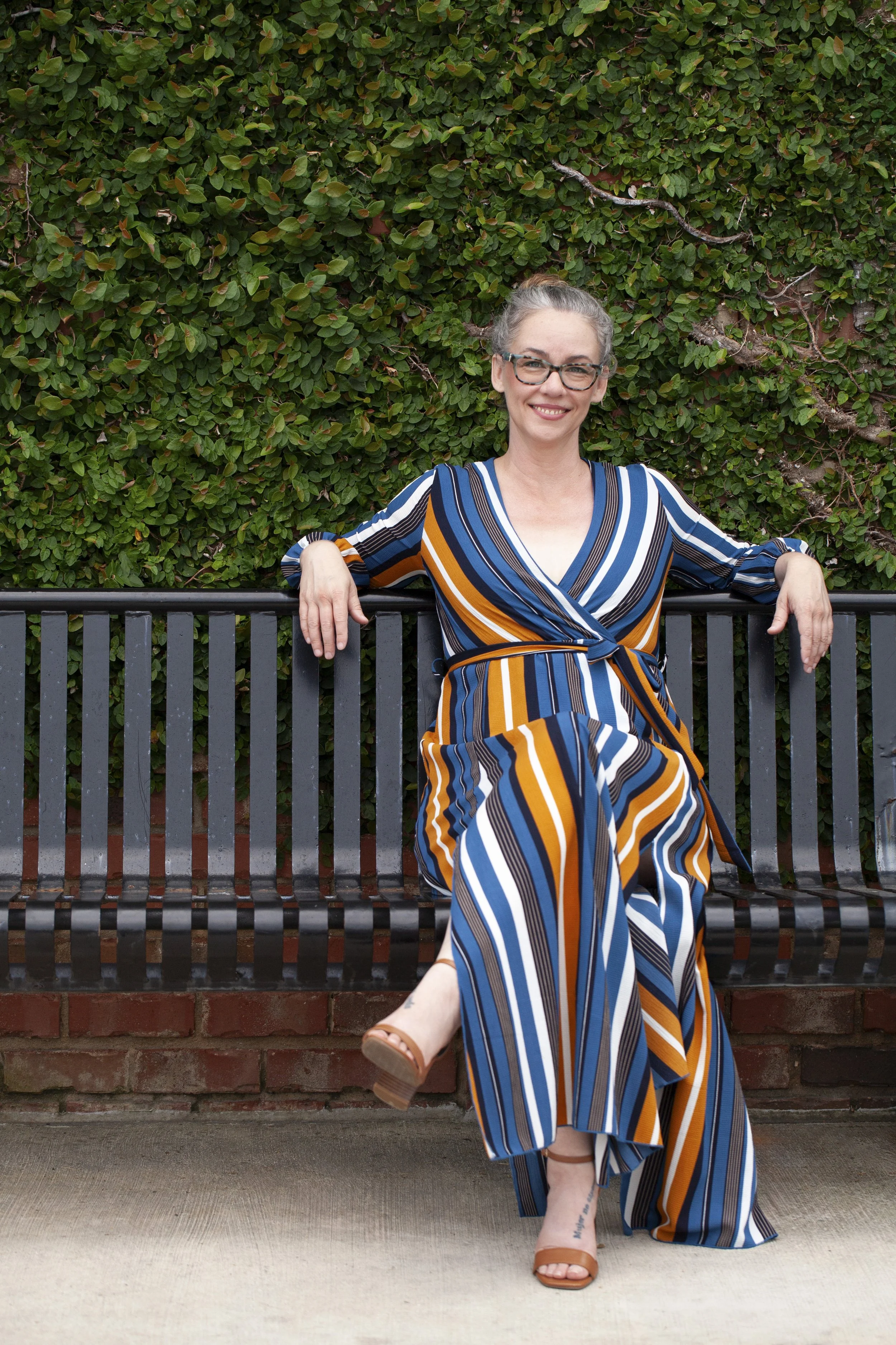 Trauma therapist smiling and leaning back on a black metal bench in front of a leafy green wall, wearing a striped blue, orange, and white wrap dress with tan sandals, one leg crossed casually in front.
