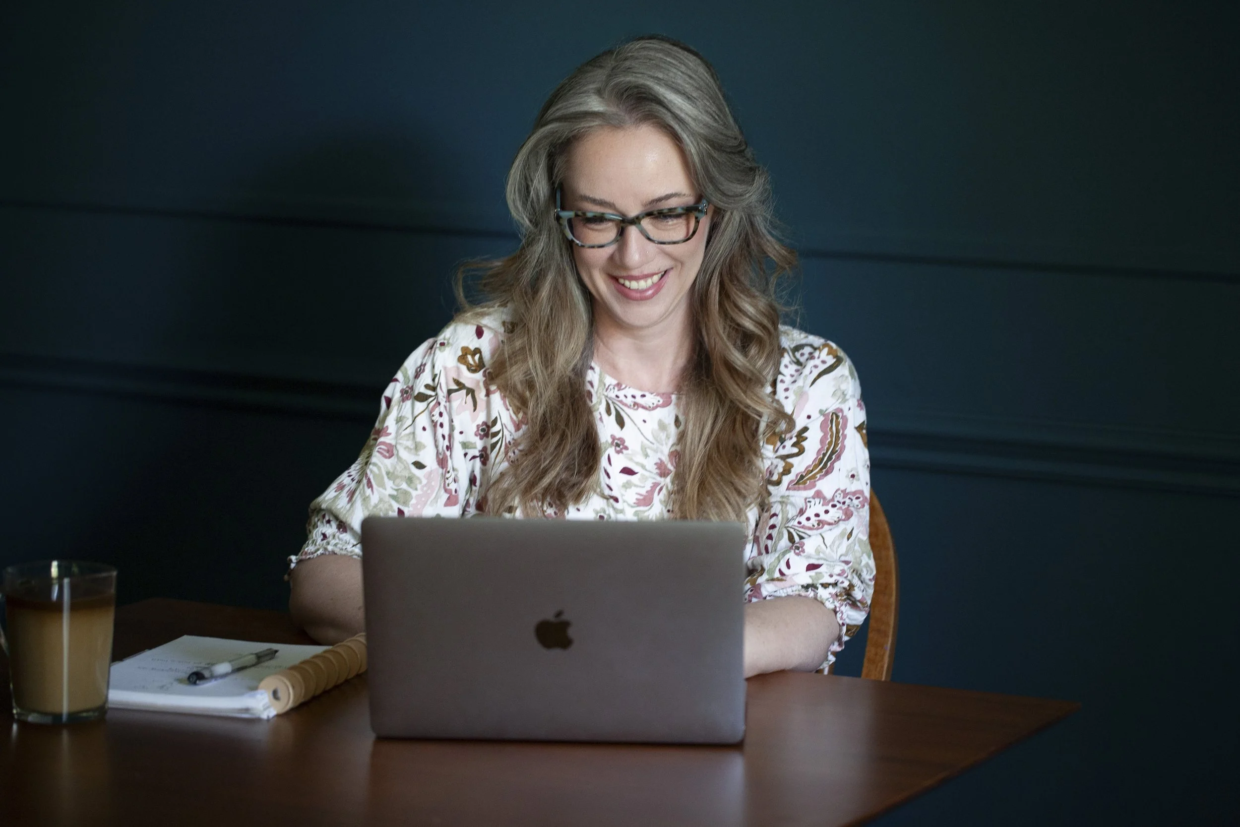 A woman with long, wavy gray hair wearing glasses and a floral blouse, smiling while working on a silver laptop at a wooden table in a dark room.