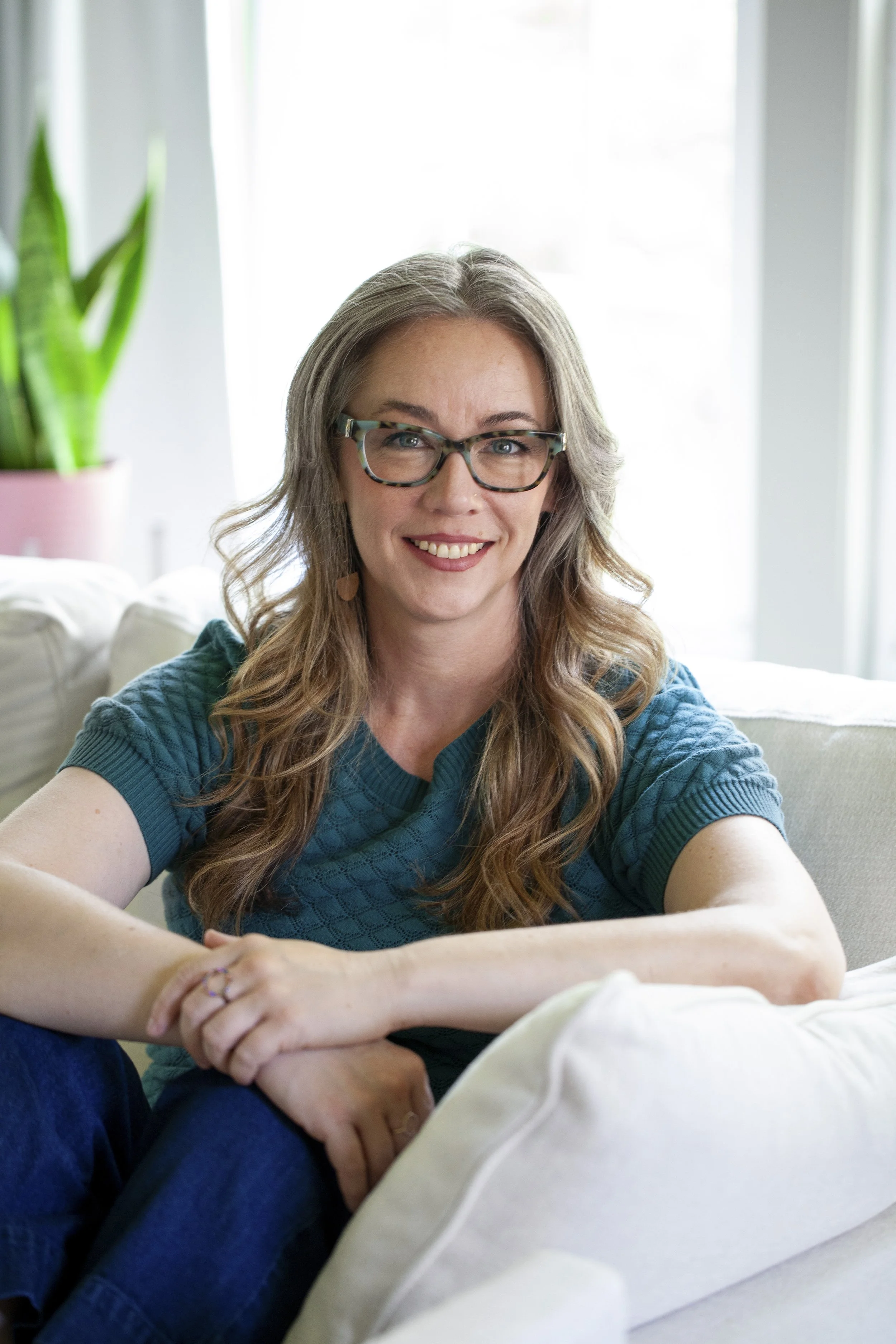 Trauma therapist smiling and sitting on a white couch, wearing glasses and a teal top, hands clasped on knee, with a potted plant and bright window light in the background.