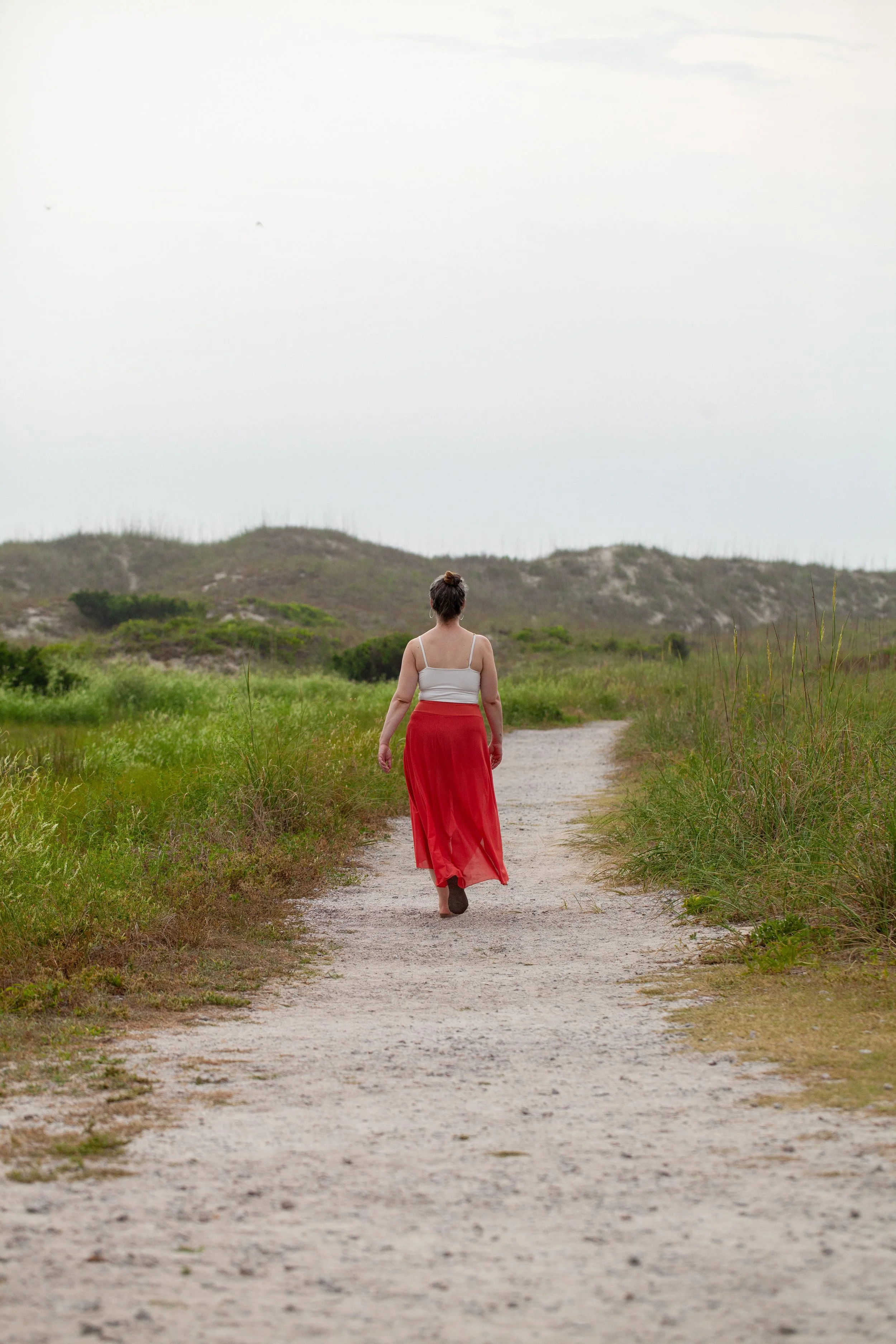 Woman seen from behind walking barefoot down a sandy path in a red maxi skirt and white top, surrounded by tall grass with dunes in the distance.
