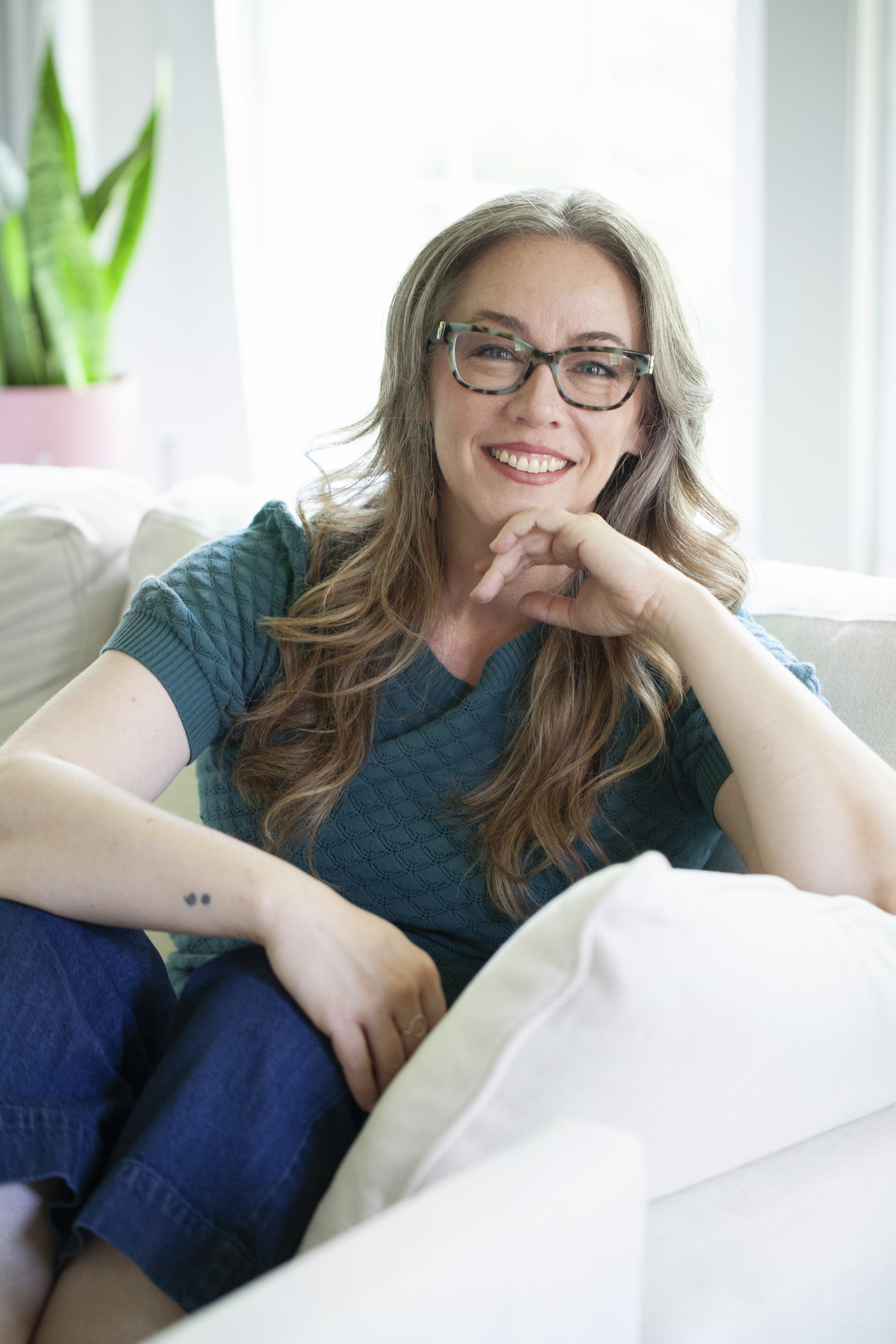 A woman with long gray hair, glasses, and freckles smiling while sitting on a couch in a bright room, wearing a teal short-sleeve shirt.
