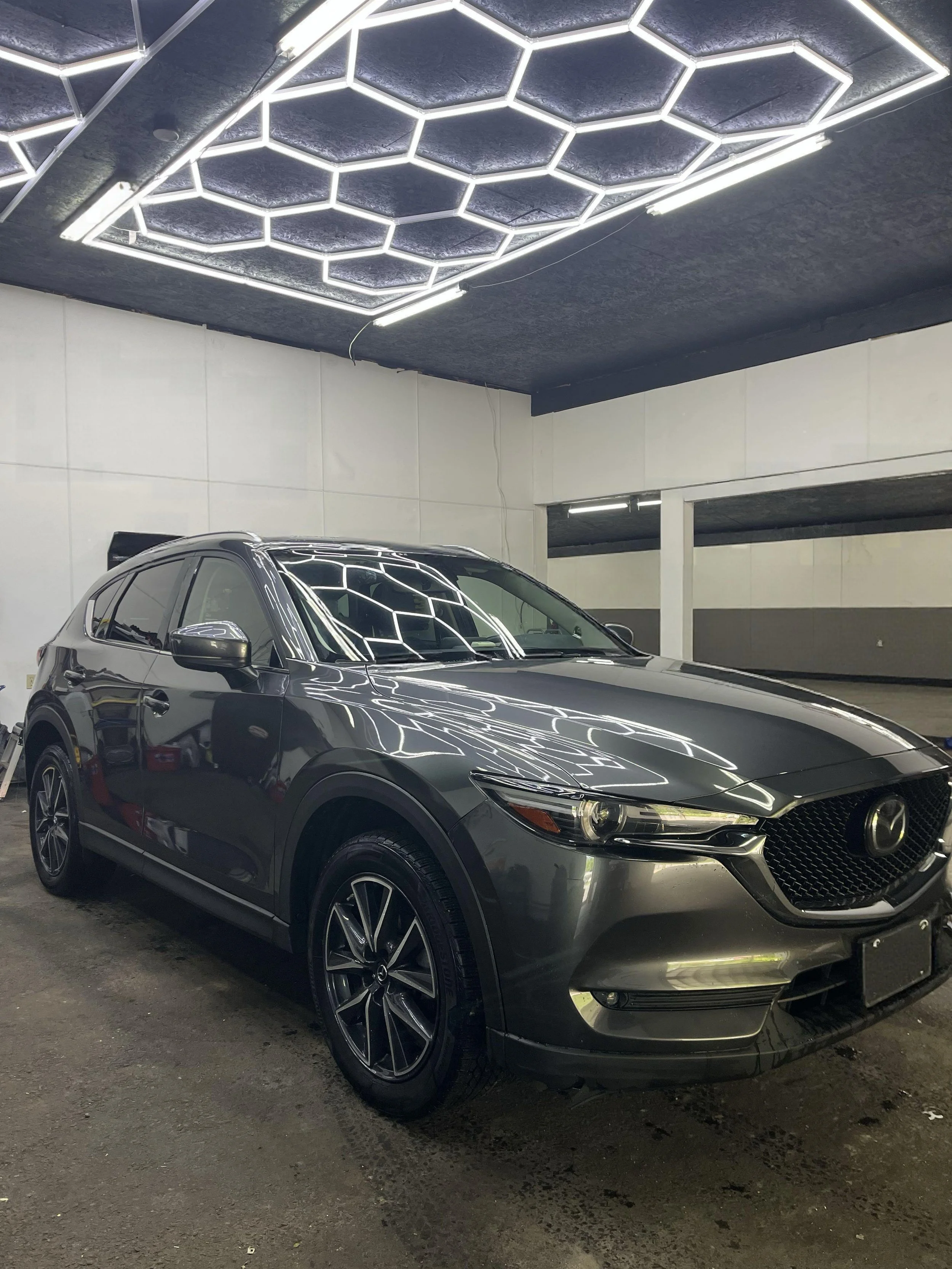A black or dark gray modern car parked in an indoor parking garage with hexagonal lighting fixtures on the ceiling, reflecting on the car's surface.