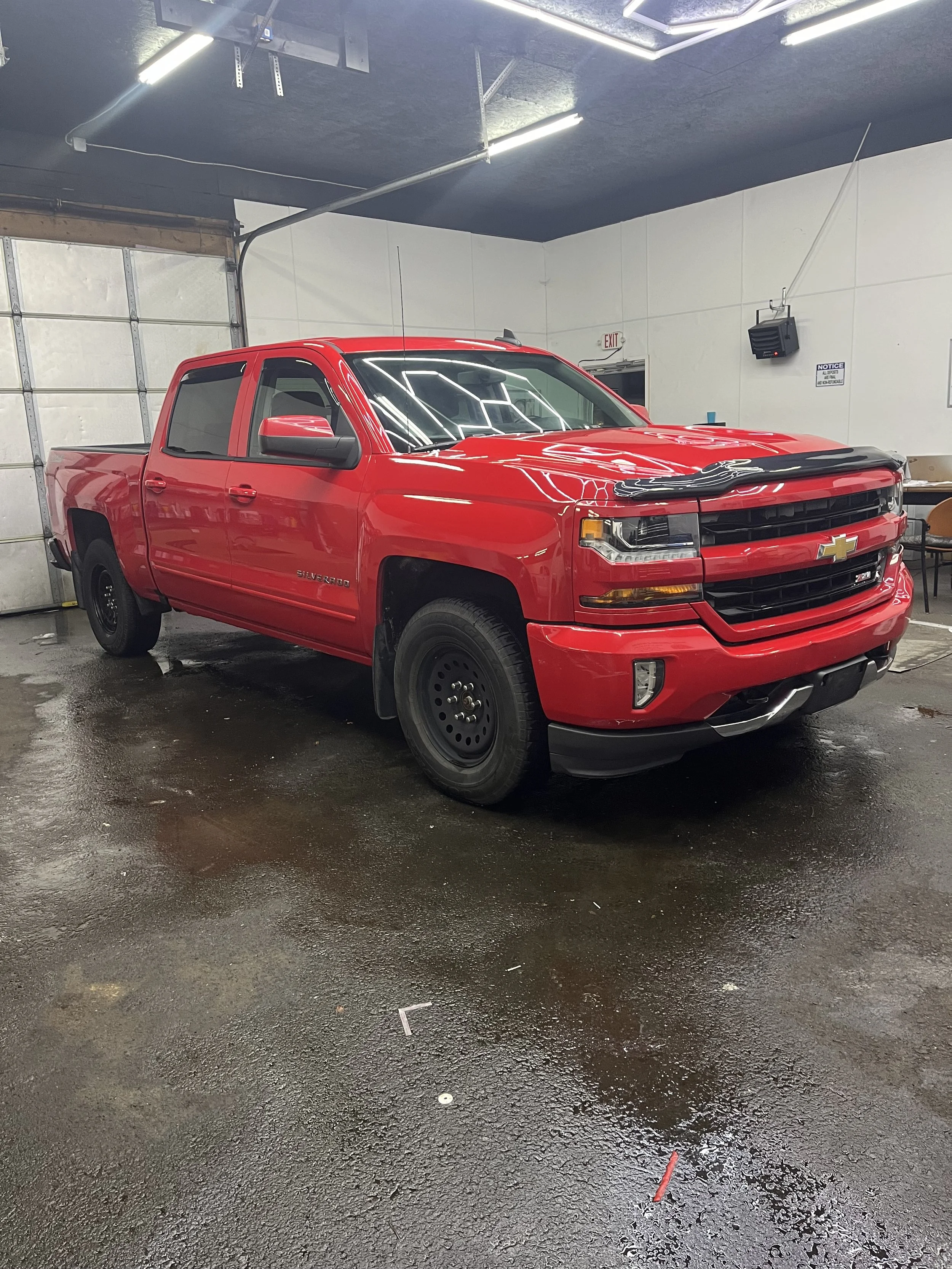 A red Chevrolet Silverado truck parked inside a garage with a concrete floor and white walls, turned slightly to the right. The garage has a black ceiling with fluorescent lighting and a partially visible garage door in the background.