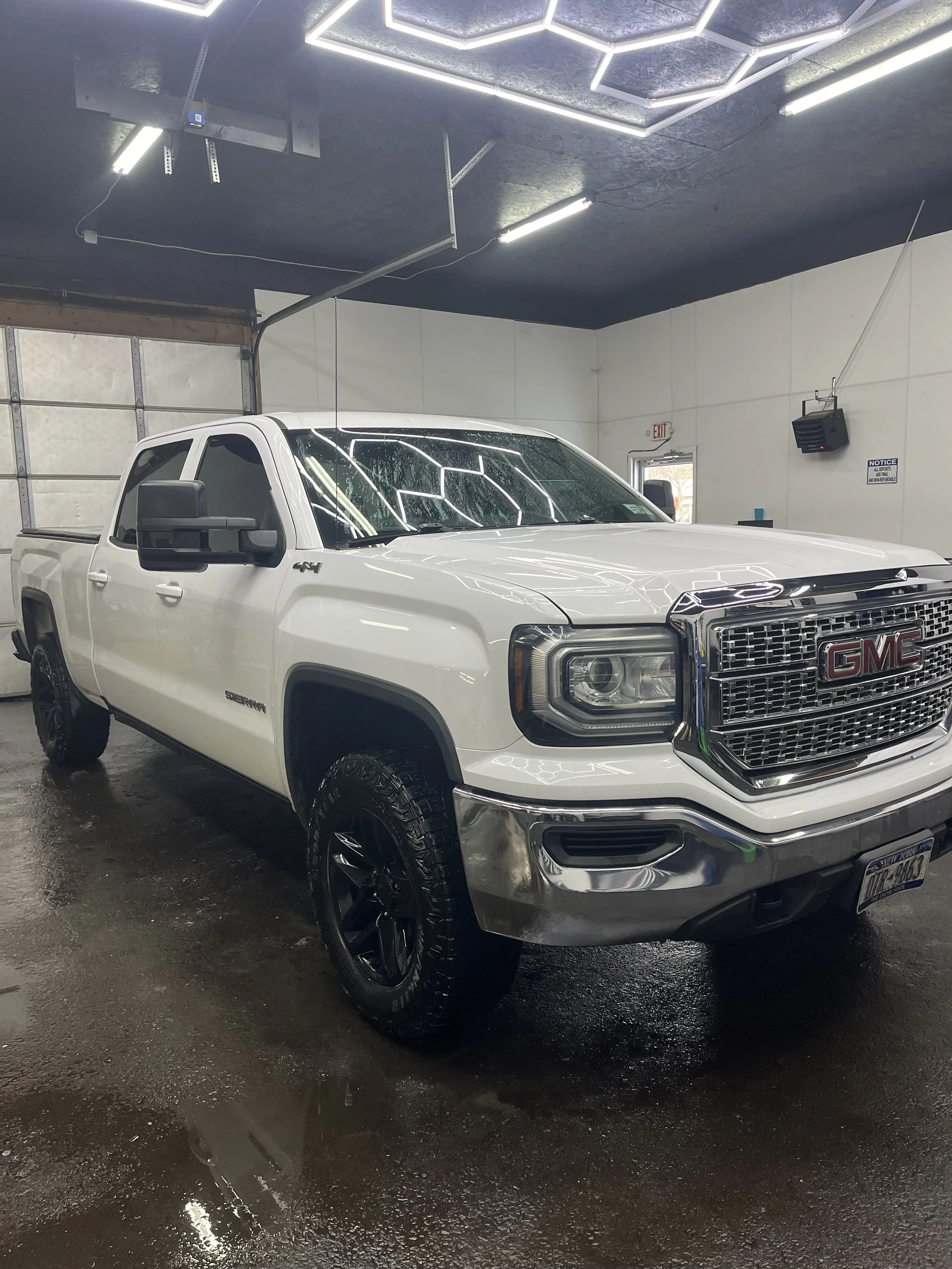 White GMC pickup truck inside a garage with a black ceiling and bright overhead LED lights, wet floor, and a garage door in the background.