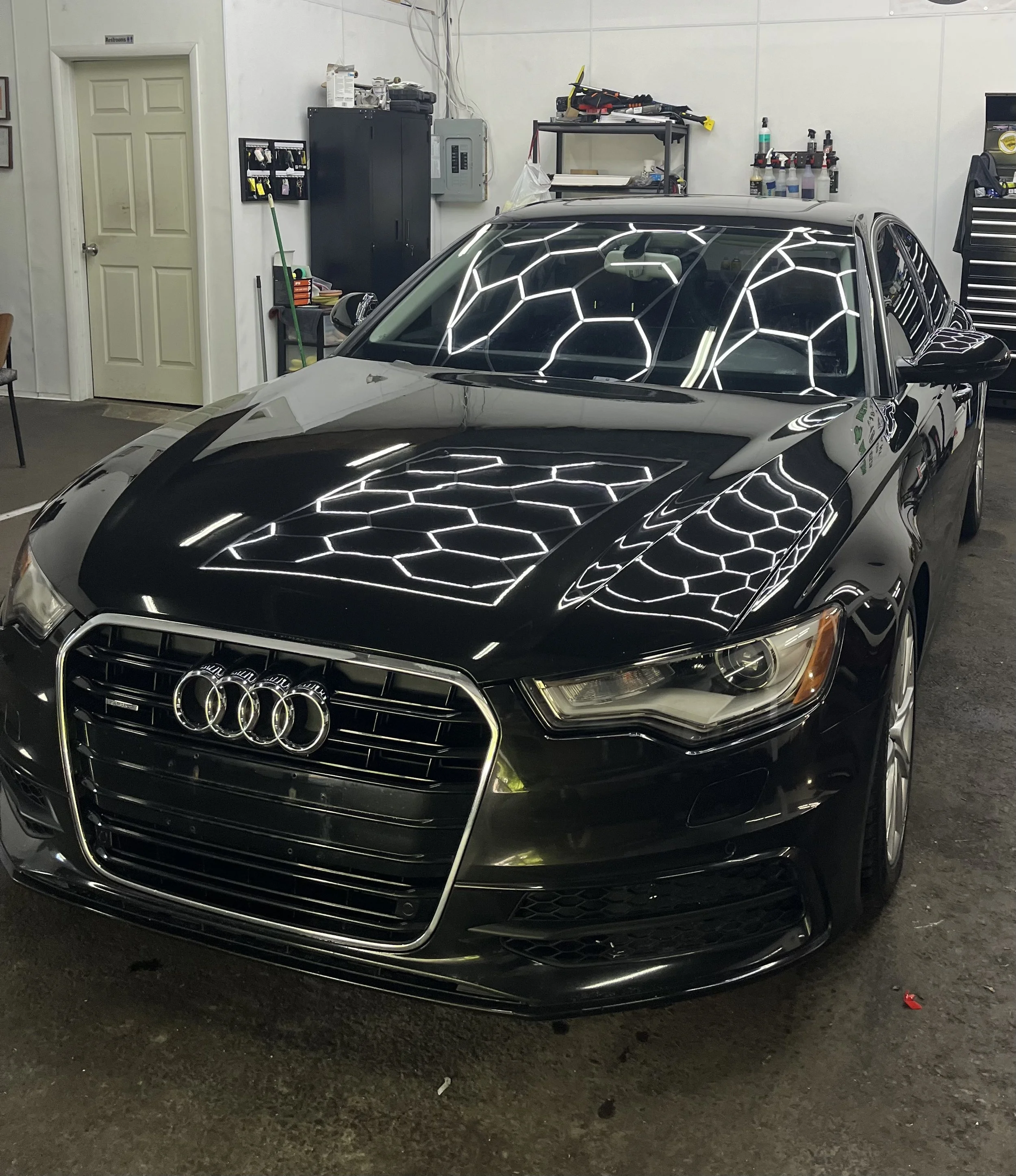 A black luxury car parked inside a garage with reflections of hexagon-shaped ceiling lights on its hood and windshield. The garage has white walls, shelves with supplies, a black metal cabinet, a broom, and various tools and bottles on the wall.