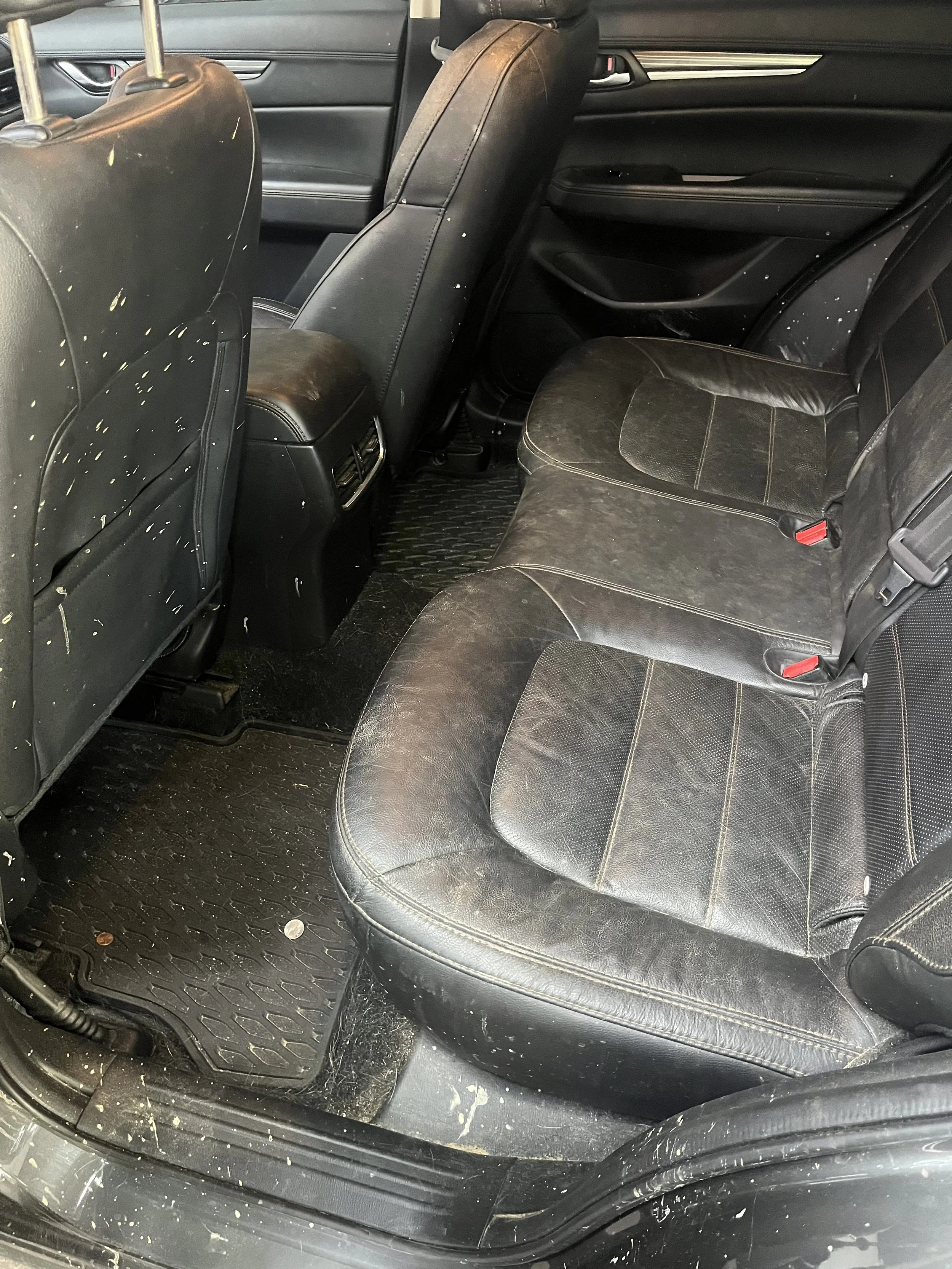 Backseat area of a car with black leather seats, some paint splatters on the seats and floor, and floor mats.