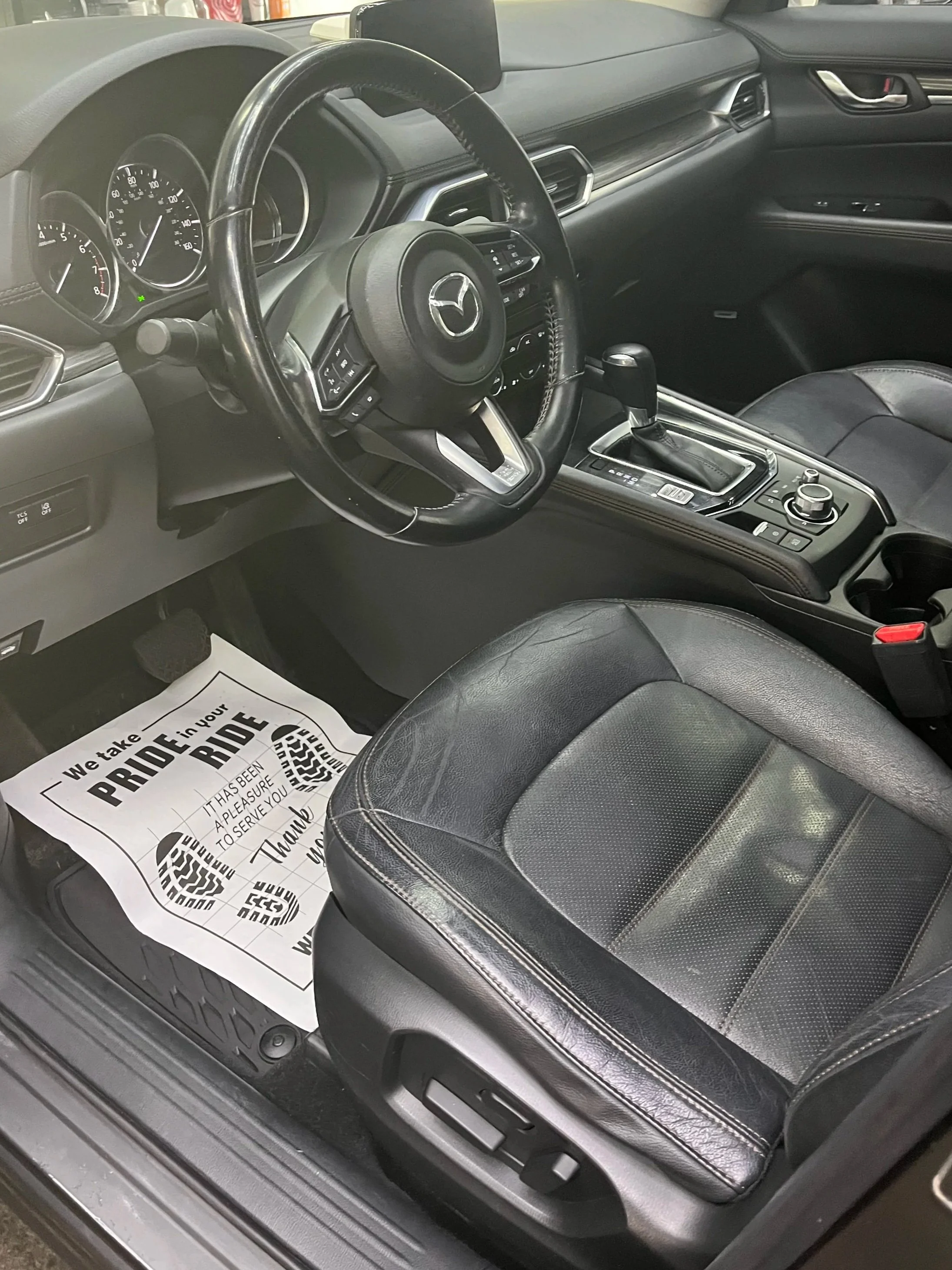 The interior of a black car, showing the driver's side with a steering wheel, dashboard, gear shift, and a black leather seat. There is a paper floor mat with a message that says "We take pride in your ride" on the floor.