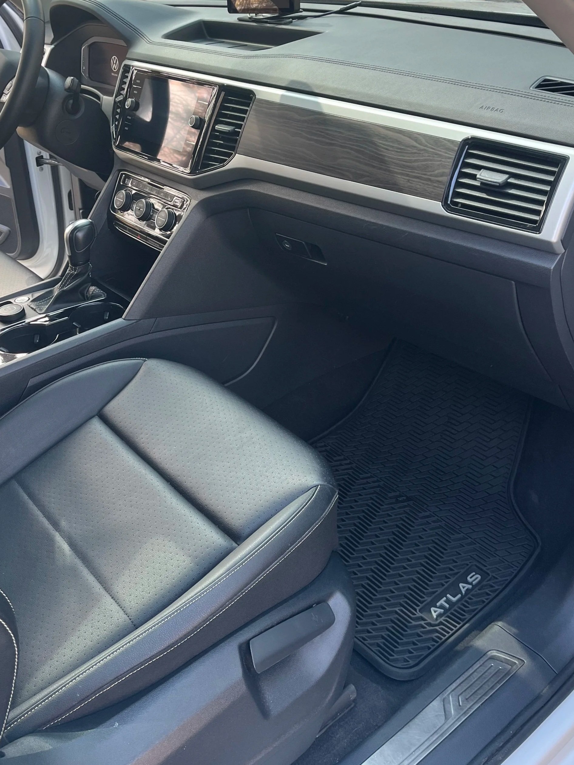 Interior of a Volkswagen Atlas vehicle showing the front passenger seat, dashboard with wood trim, center console, gear shift, and floor mat with 'Atlas' branding.