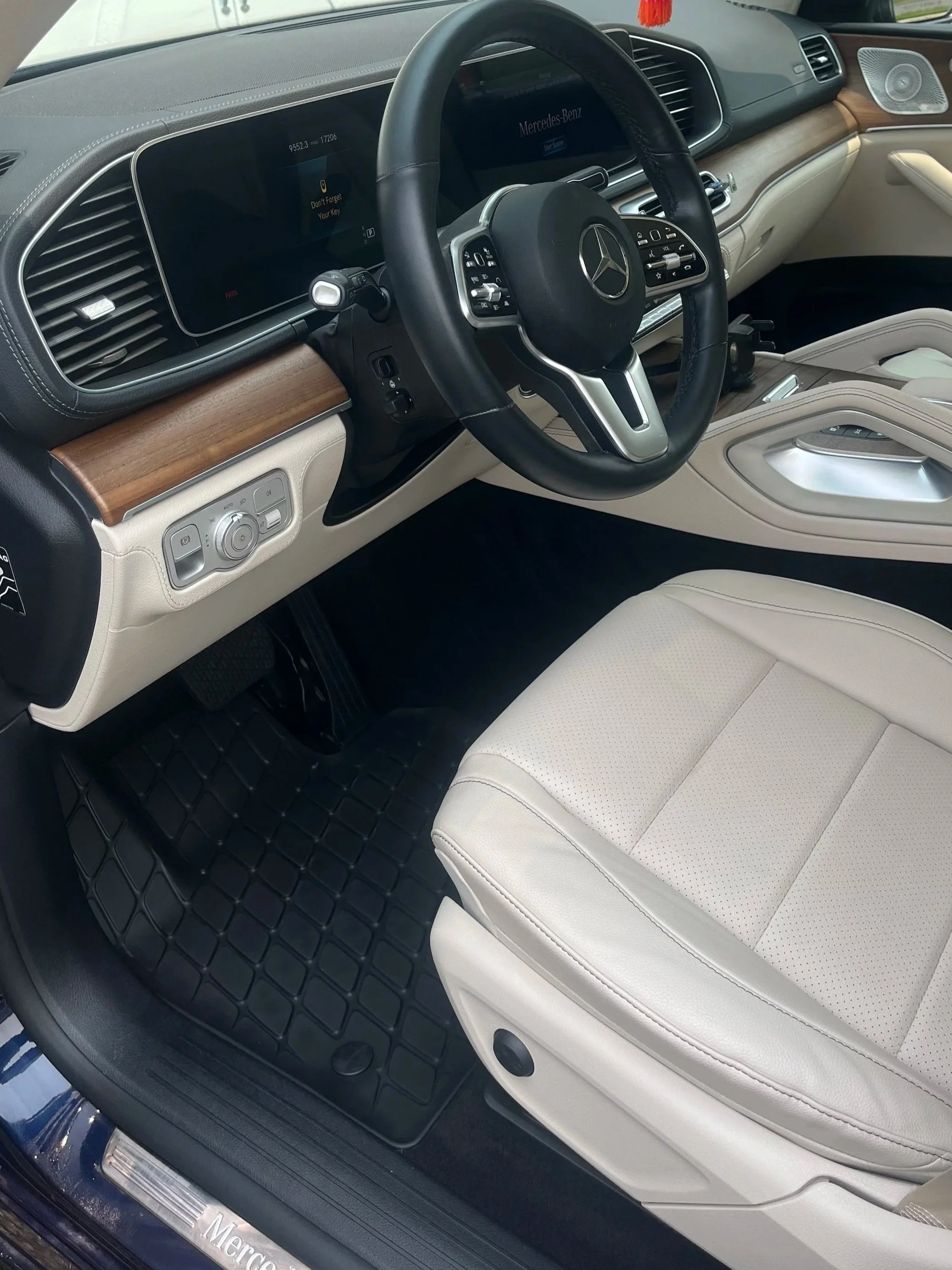 Interior of a luxury Mercedes-Benz car with a black steering wheel, digital dashboard, and beige seats, floor mat and door trim, featuring wood accents and modern control buttons.