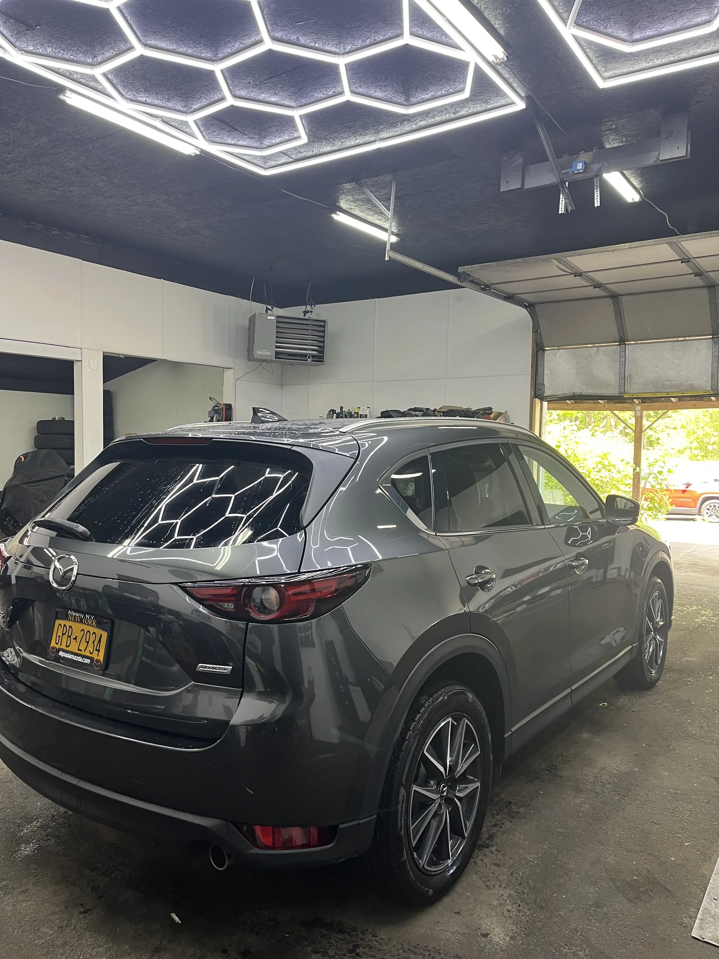 A silver Mazda SUV parked inside a garage with a black ceiling featuring hexagon-shaped LED lights. The vehicle has New York license plates and there are tools and supplies on a shelf in the background.