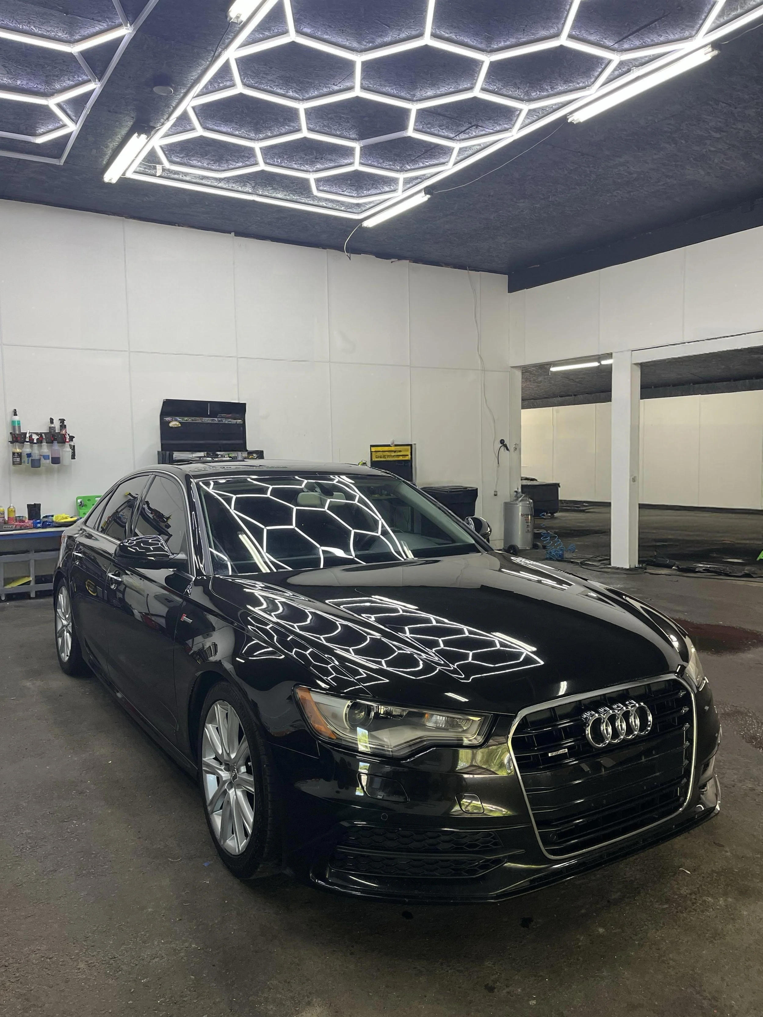Black Audi car parked inside a garage with hexagonal light fixtures on the ceiling reflecting on the car's surface.