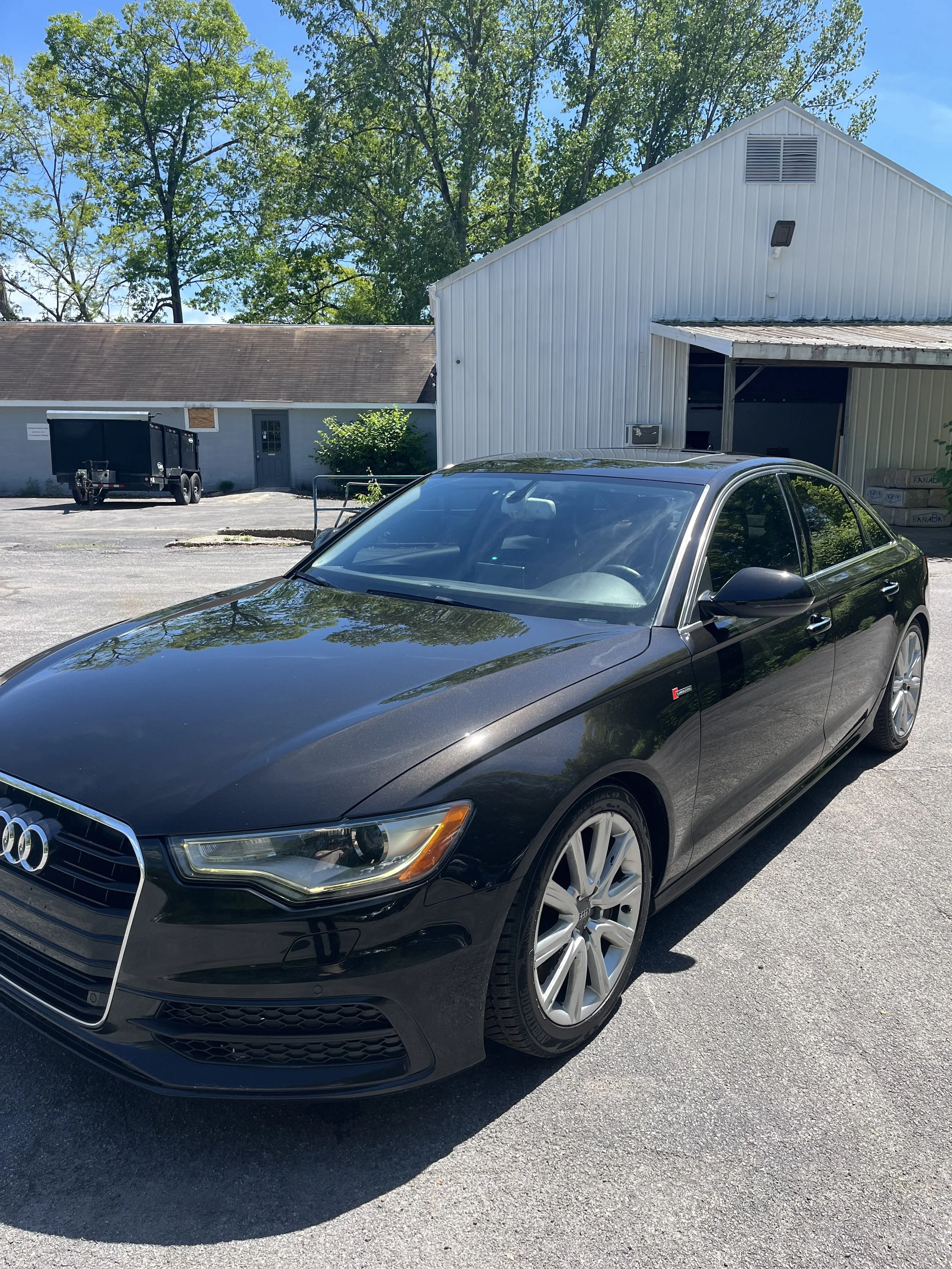 A black Audi sedan parked on a paved lot in front of a white building with a metal roof, adjacent to a smaller gray building and surrounded by green trees under a clear blue sky.