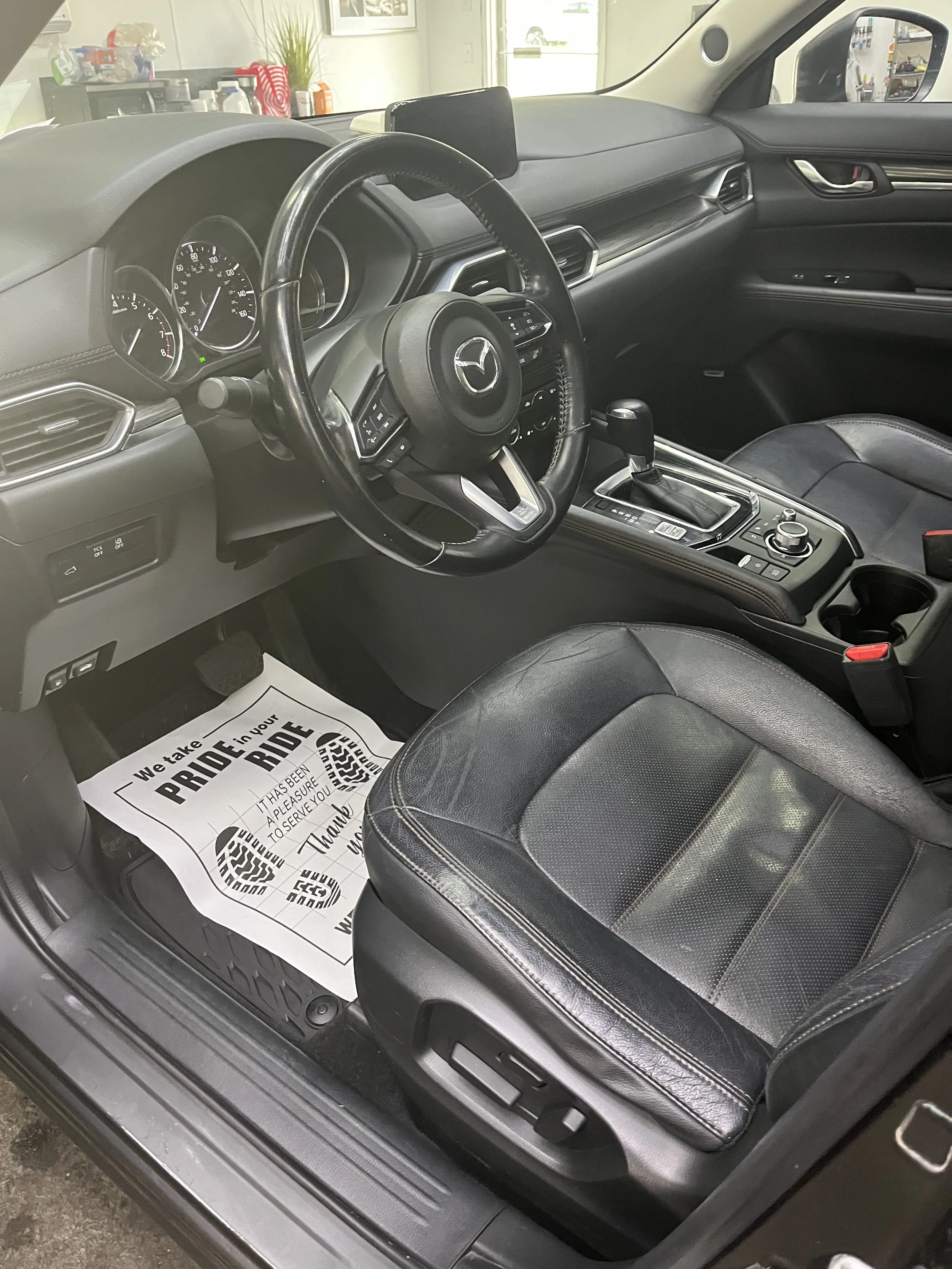 The interior of a Mercedes-Benz vehicle showing the steering wheel with a Mercedes logo, the dashboard with gauges, the gear shift lever, and a black leather seat. There is a paper floor mat with printed text on the driver's side floor.