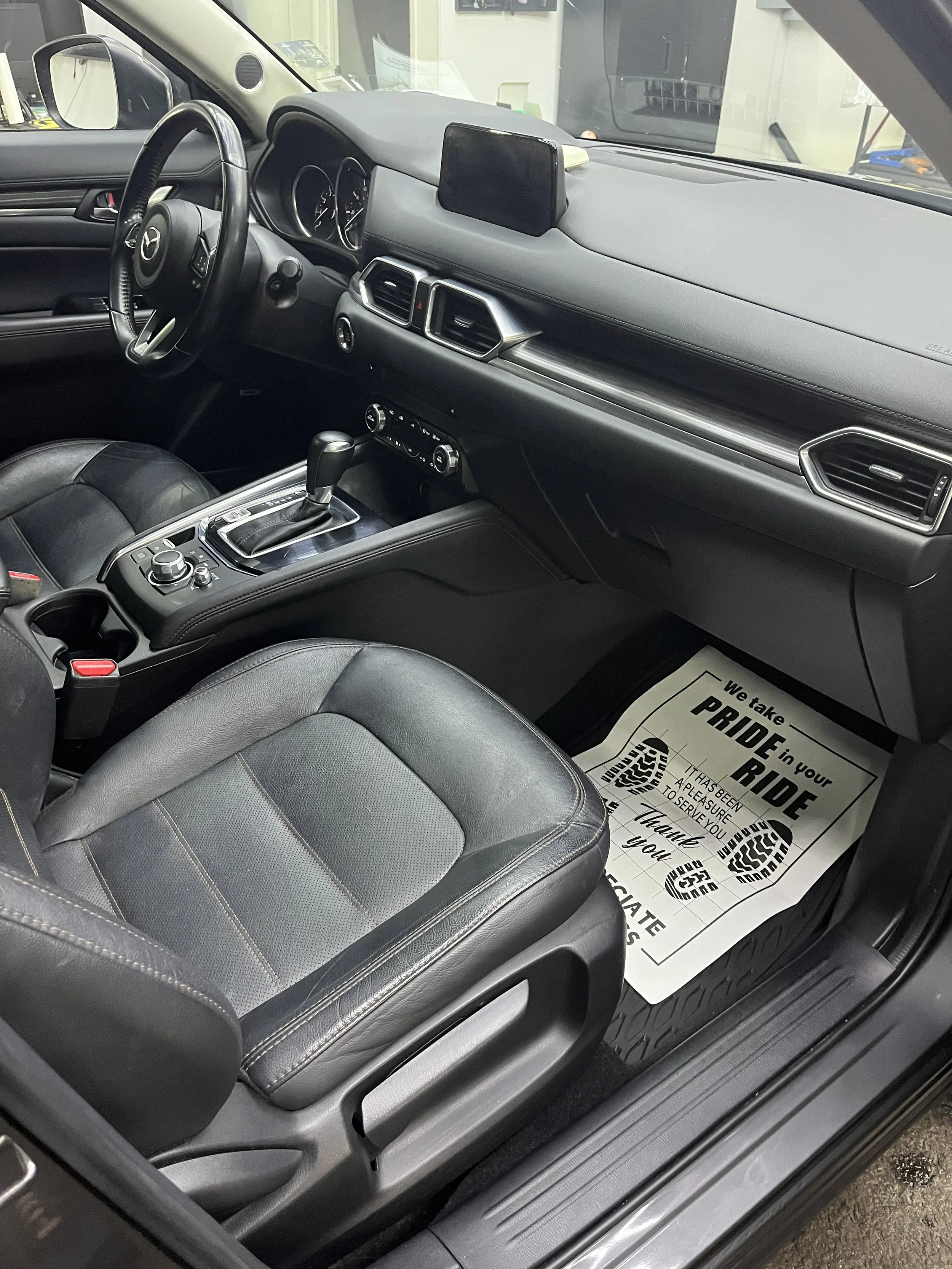 The interior of a Mazda vehicle showing the driver's seat, dashboard, steering wheel, center console, and a paper floor mat with a message about taking pride and service, with footprints printed on it.