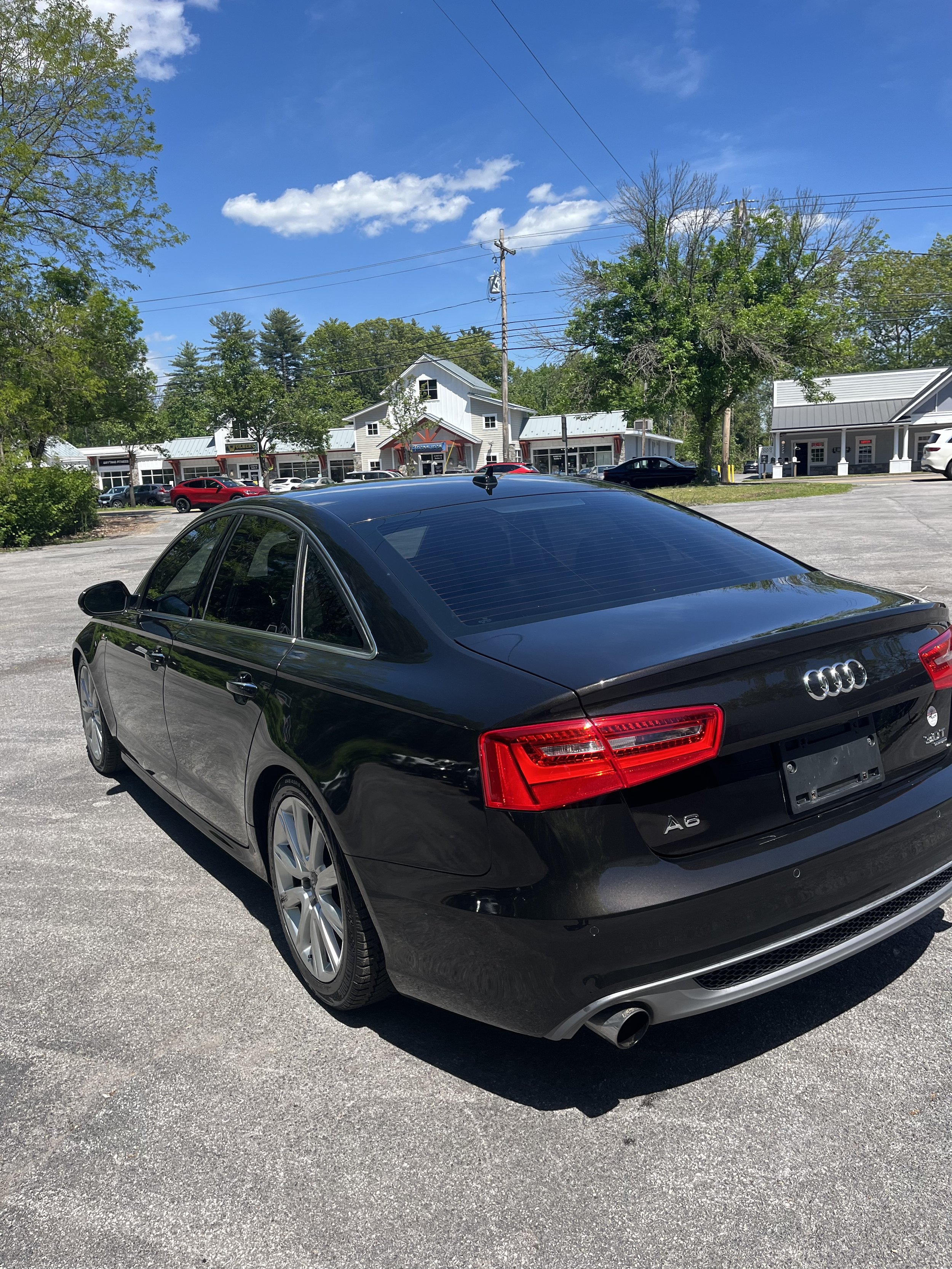 A black Audi A6 parked in a lot with storefronts and trees in the background under a partly cloudy sky.
