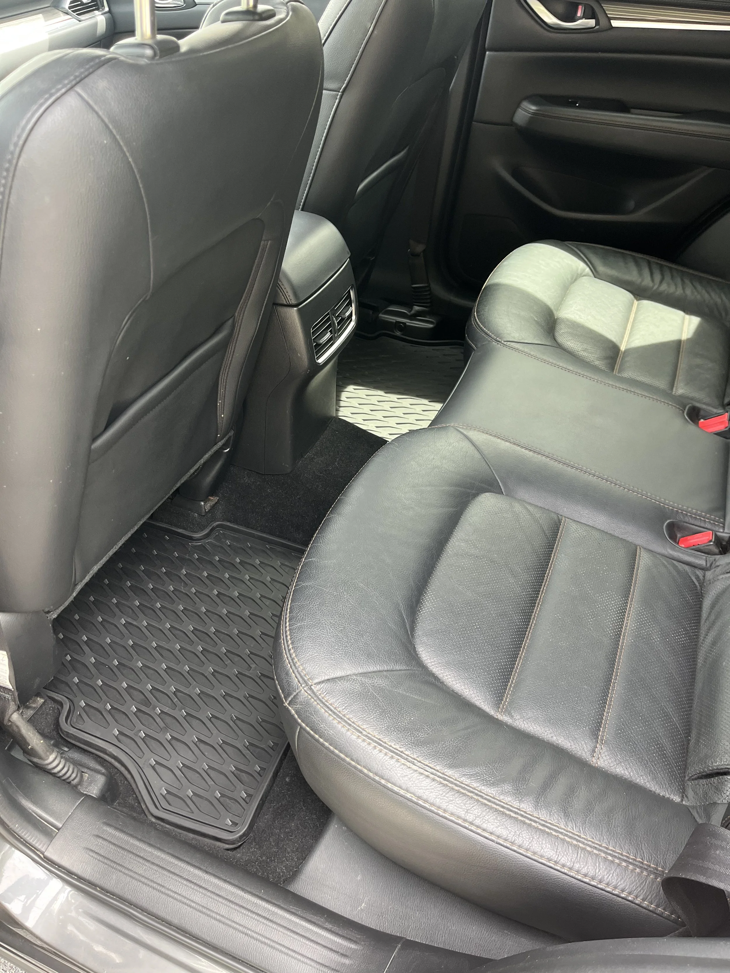 The image shows the interior of a car focusing on the back seat and floor. The back of the front seats is visible, along with an air vent and part of the center console. The floor has a black rubber mat with a textured pattern.