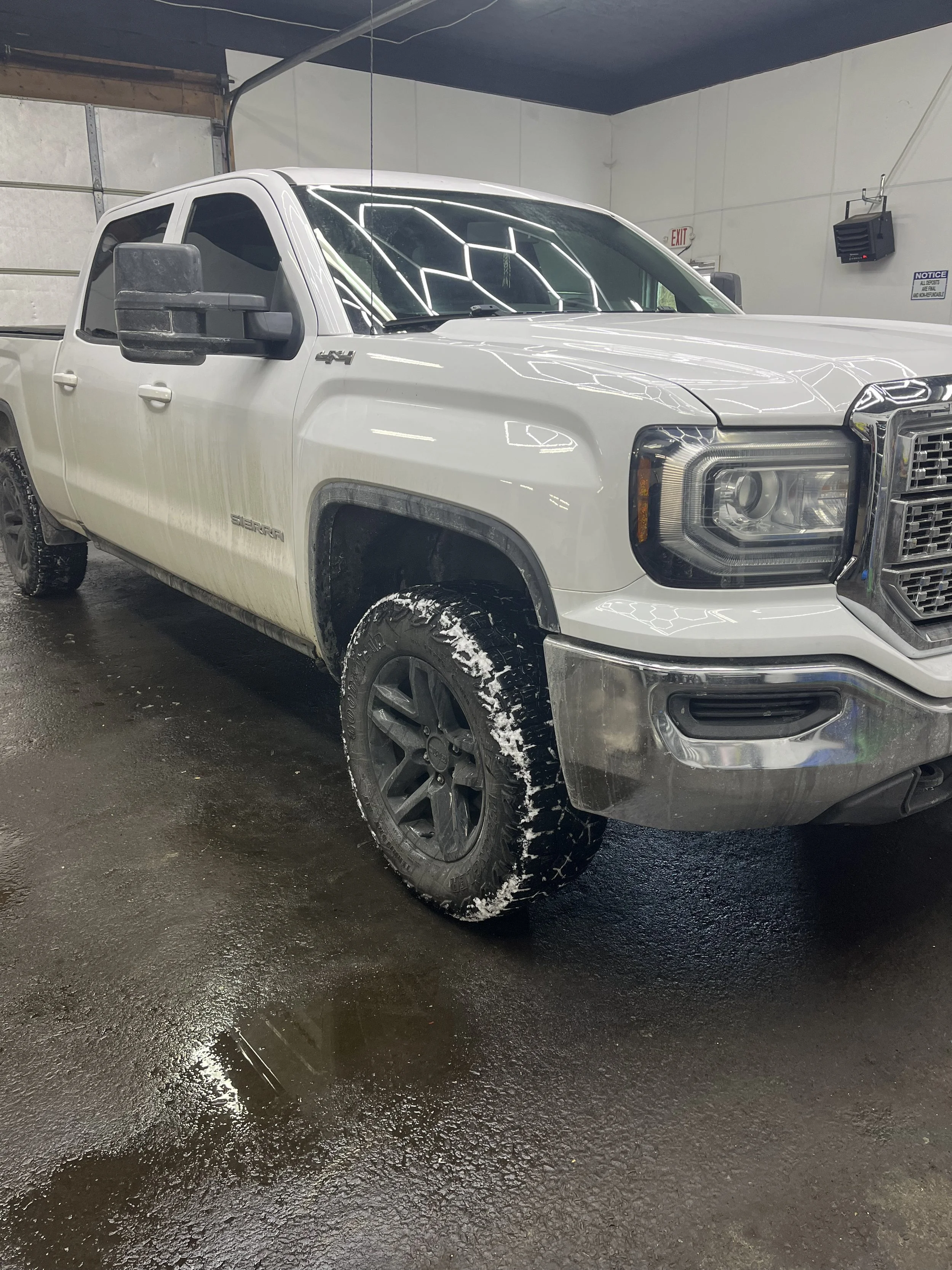 A white GMC Sierra pickup truck with black wheels and side mirrors, parked inside a garage with wet black concrete floor, fluorescent ceiling lights and garage door in the background.