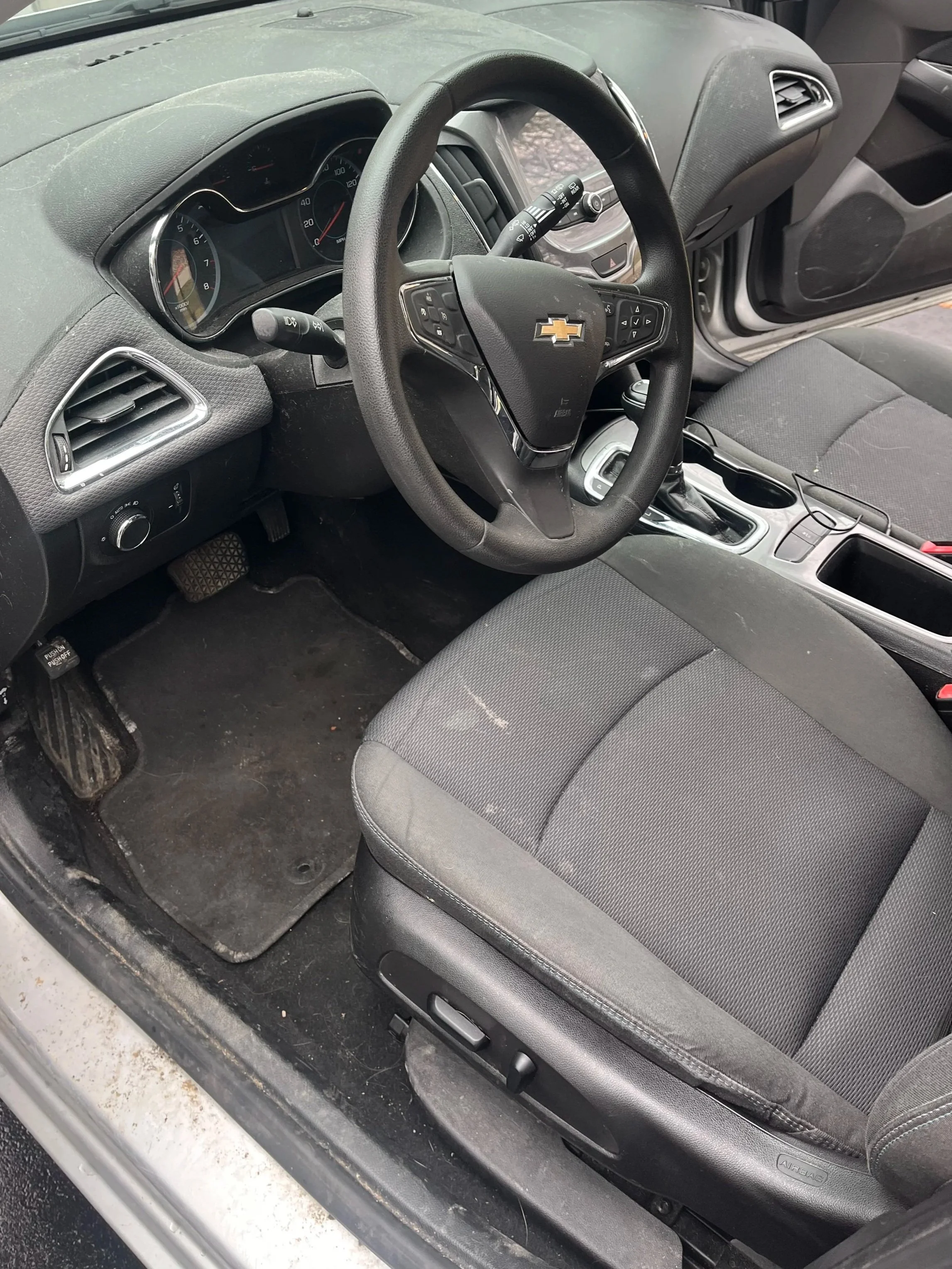 Inside view of a car's front passenger area showing the driver's seat, dashboard, steering wheel with Chevrolet logo, gear shift, and floor area with a dirty pedal and floor mat.