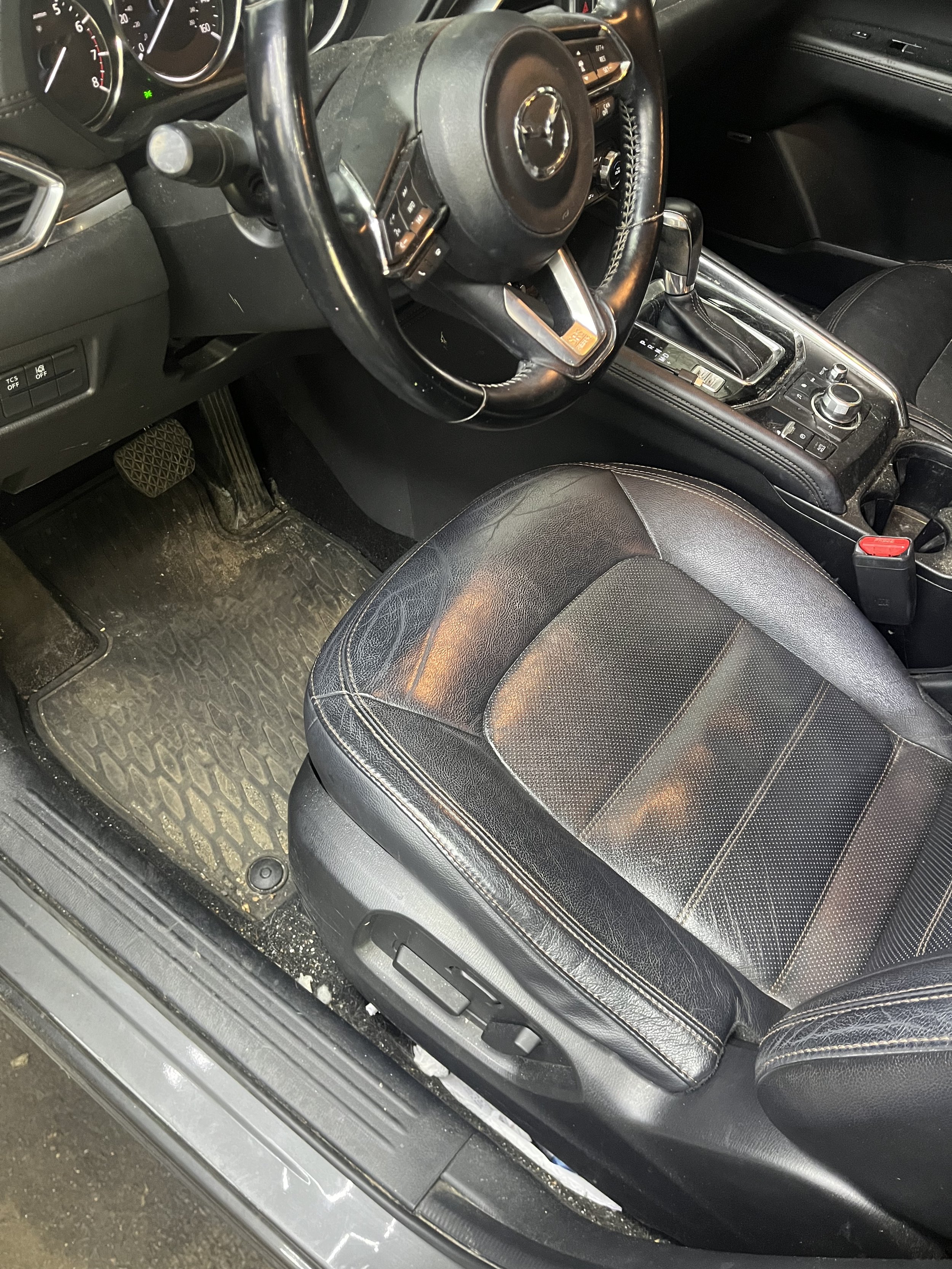 The image shows the interior of a car, including the steering wheel, gear shift, and driver's seat. The floor mat is dirty, and the dashboard features various controls and dials.