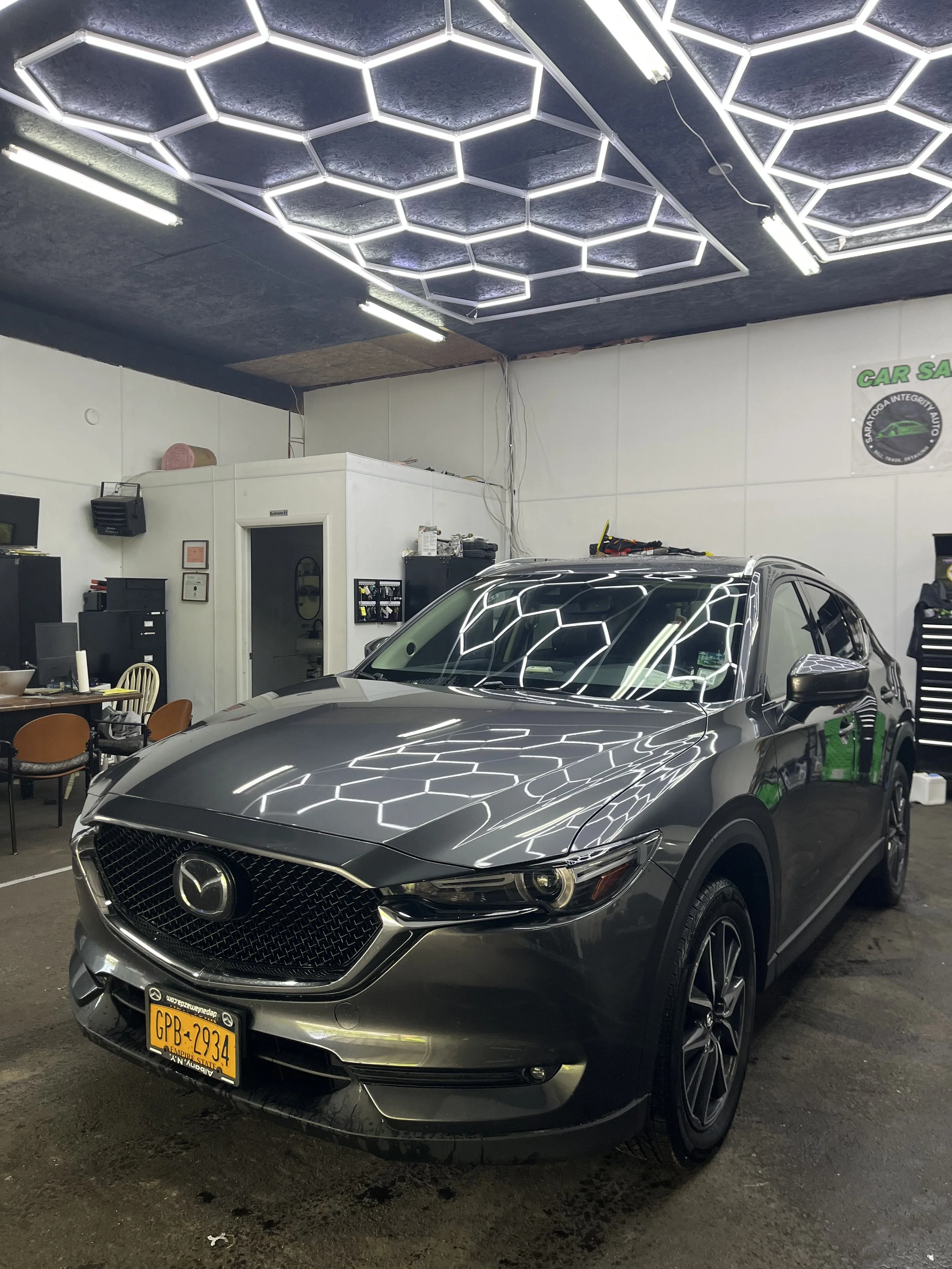 A silver Mazda SUV with New York license plates parked indoors, reflecting a hexagon-patterned LED ceiling light.