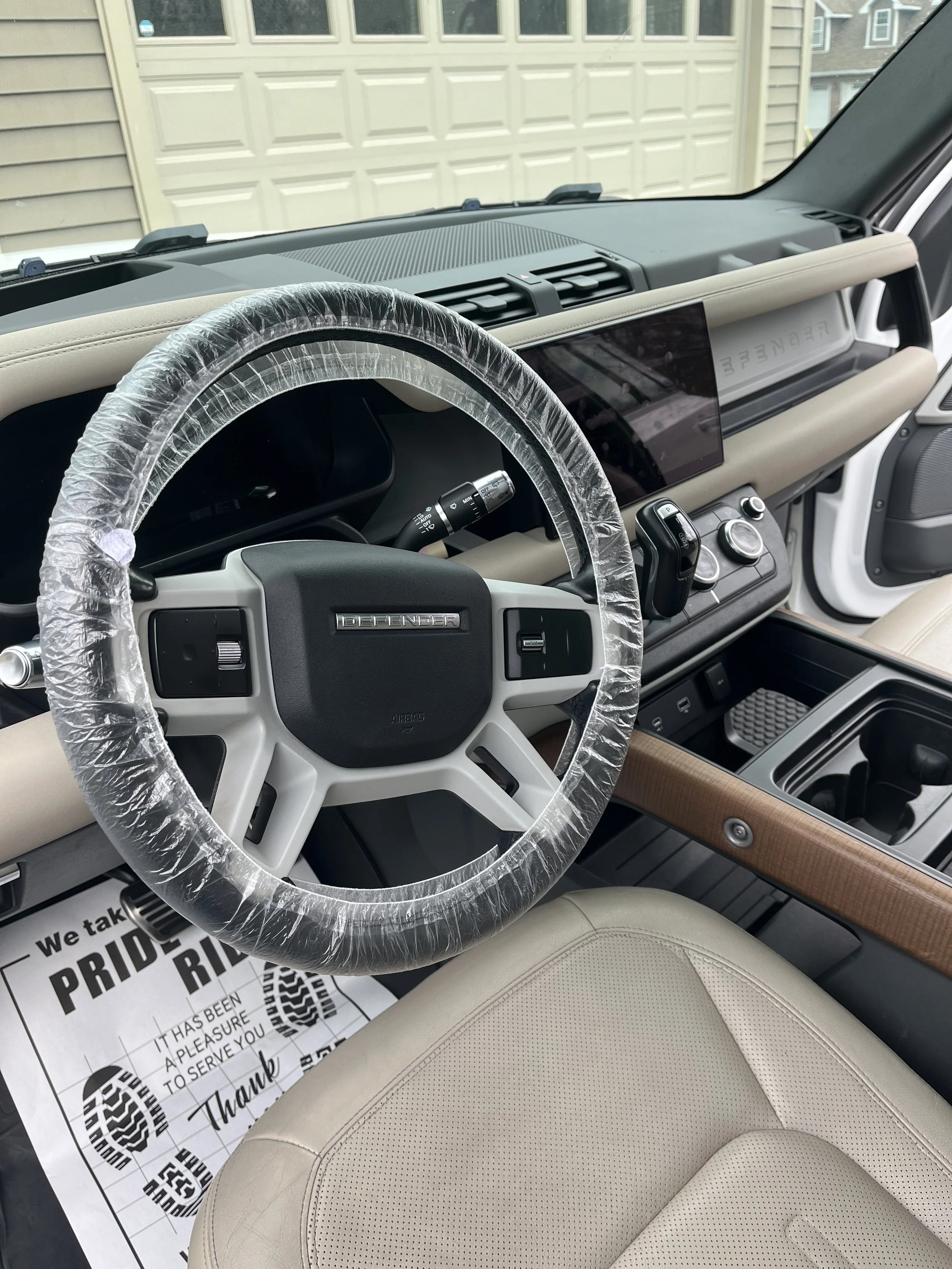 Interior of a vehicle with a beige leather seat, a plastic protective covering on the steering wheel, a digital dashboard, and a large touchscreen display. There is a sign on the floor with text supporting pride and gratitude.