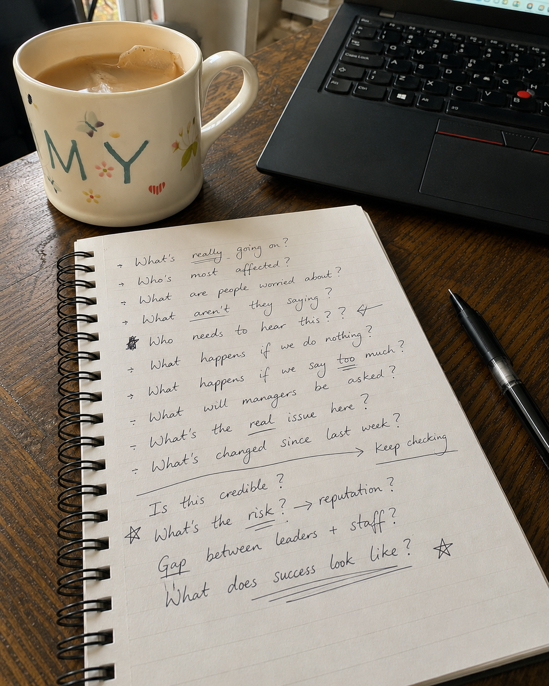 A desk scene featuring a mug with 'Mummy' written on it and the teabag left in, a black laptop, a spiral notebook with handwritten questions about communications and engagement