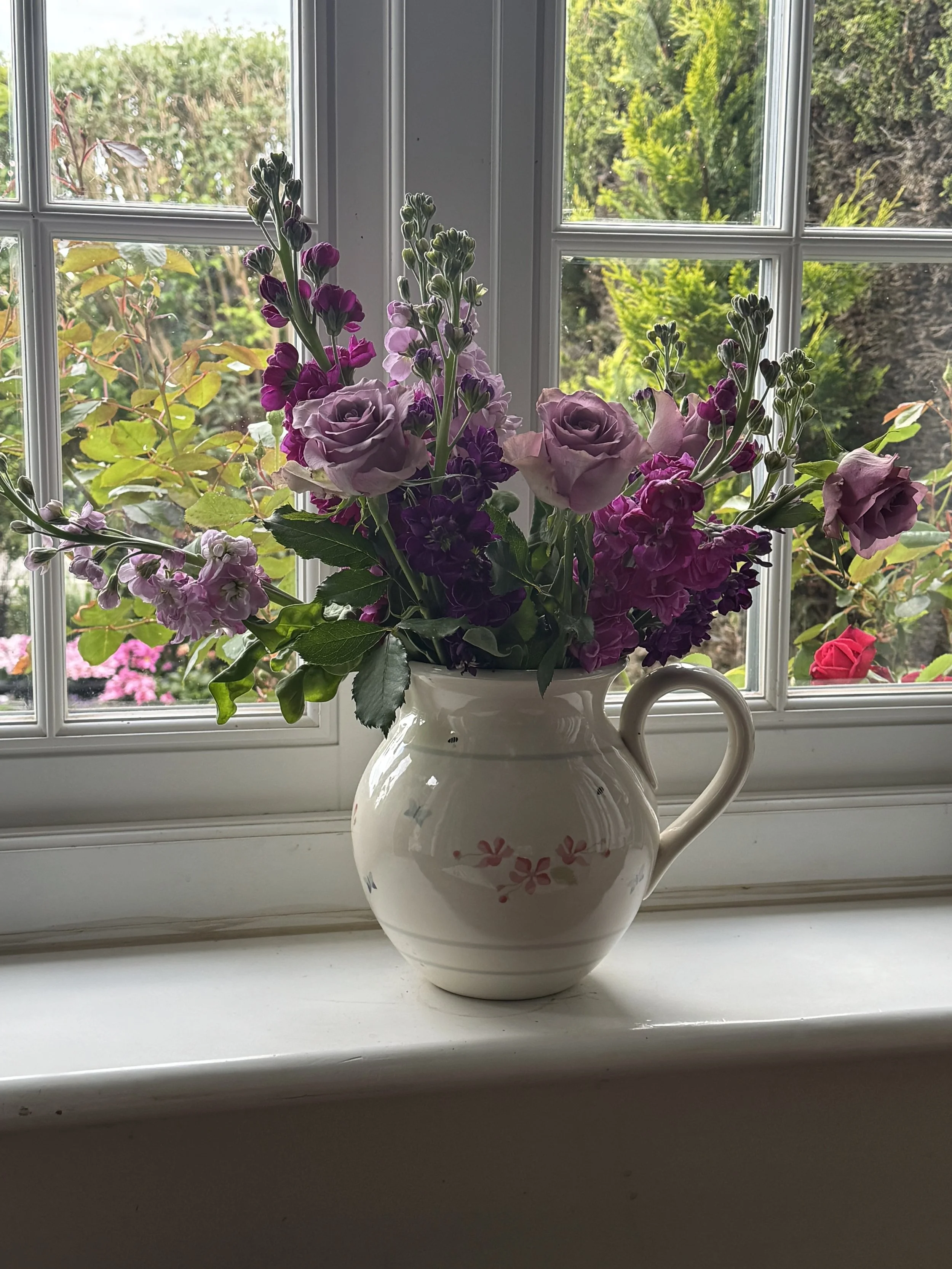 A white ceramic pitcher with pink floral patterns on a white windowsill, filled with purple, pink, and lavender flowers facing a window with an outdoor view of green bushes and trees.