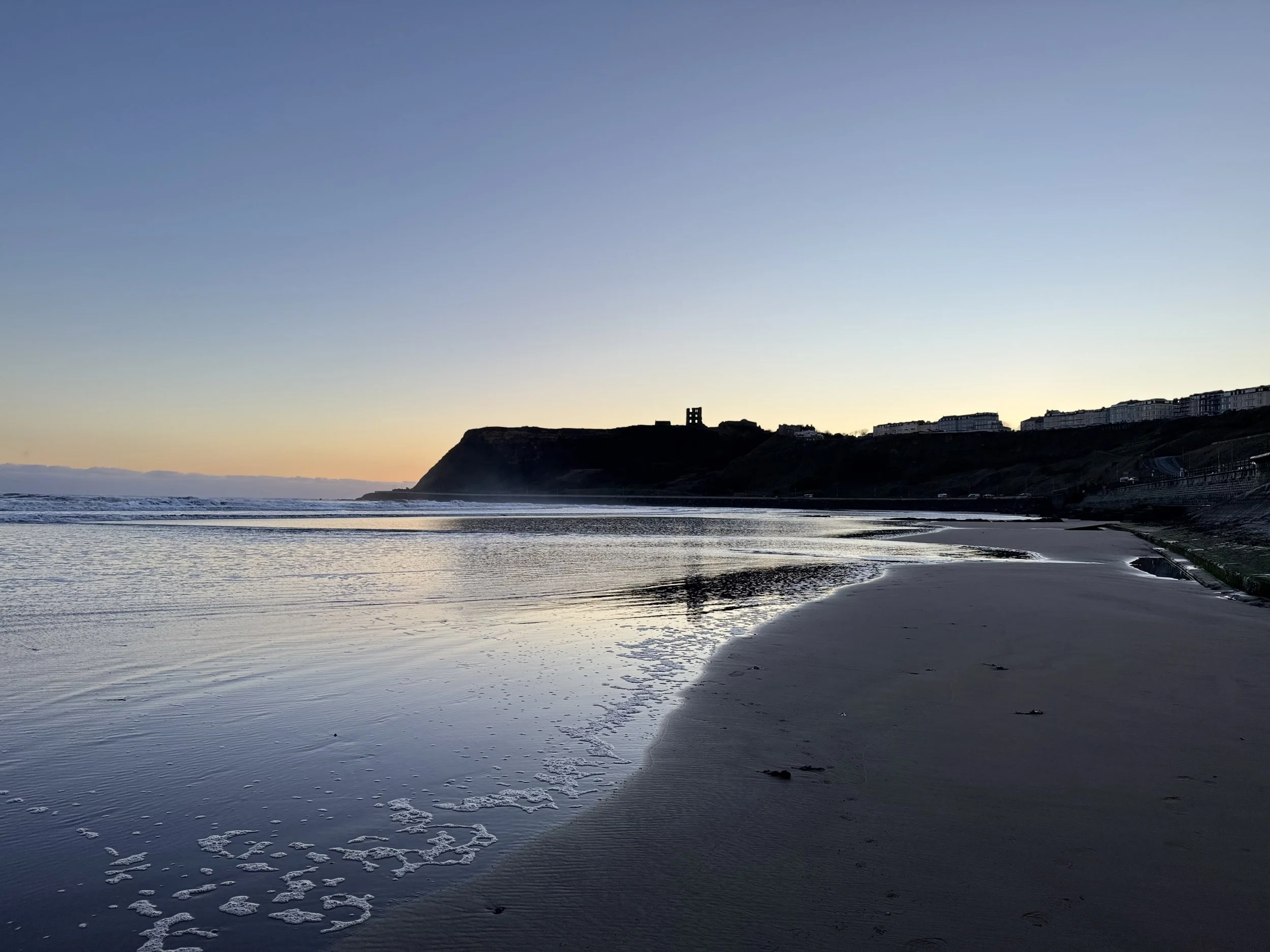Scarborough beach at sunset with footprints in the sand, calm water, cliffs with buildings, and a clear sky.