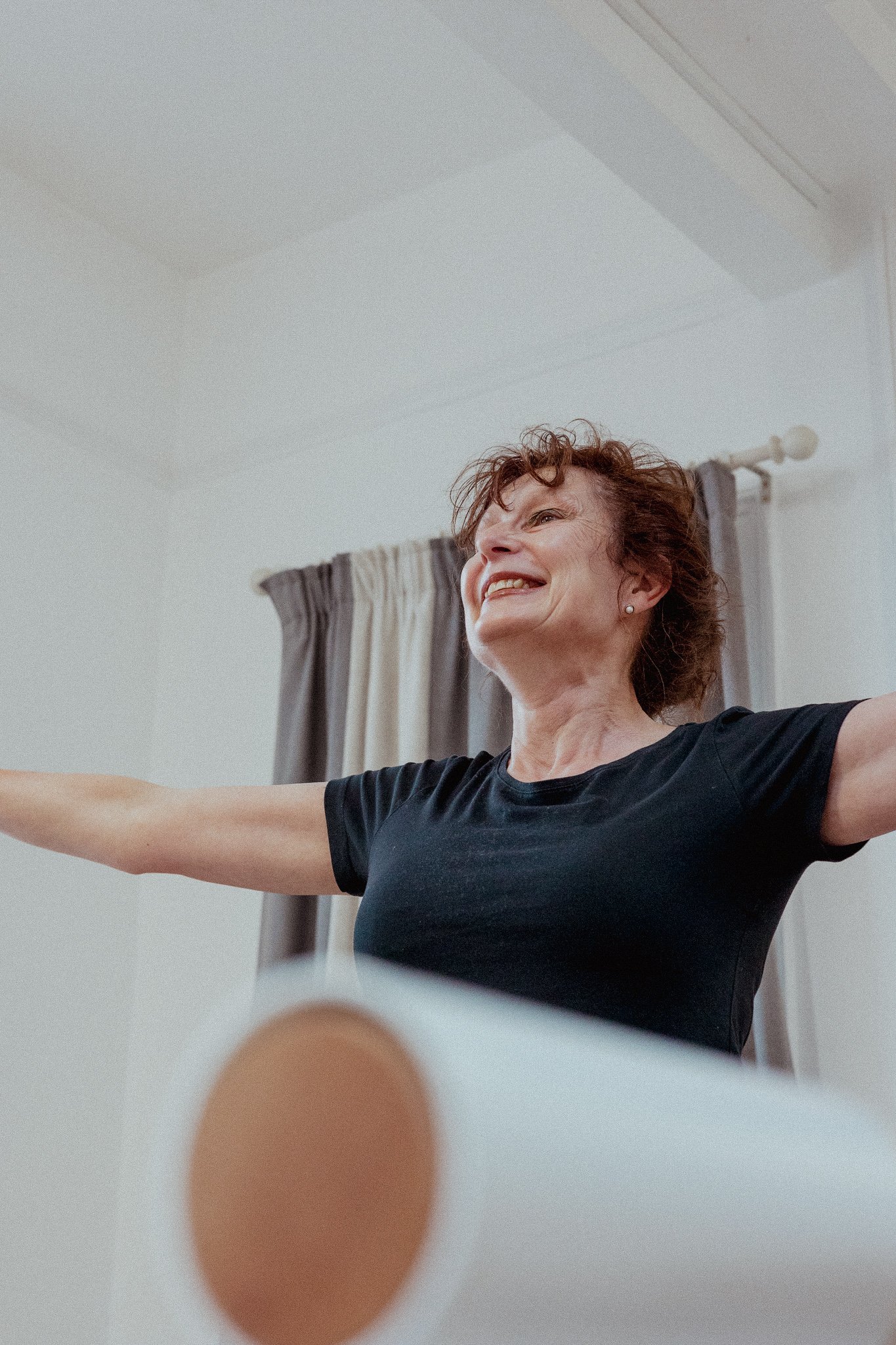 A woman with curly red hair, wearing a black shirt, extending her arms in a stretching or exercise pose in a room with curtains and a white ceiling.