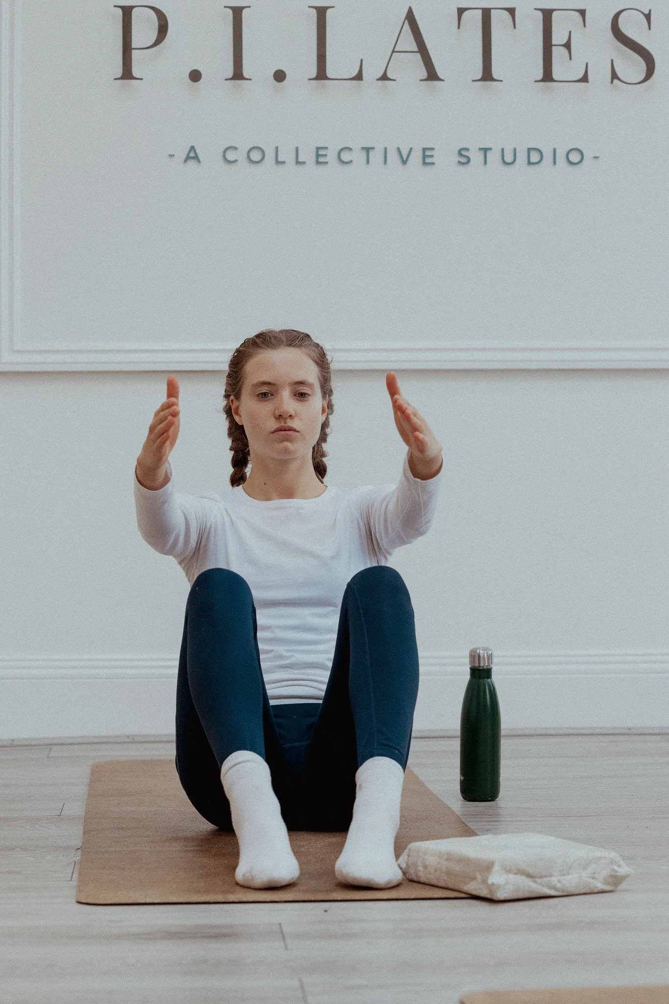 A young woman is practicing yoga or a meditation pose on a mat in a room with a sign that reads 'P.I. LATES' and 'A COLLECTIVE STUDIO'. She is sitting with her legs crossed, hands raised and facing upward, and has a focused expression on her face. There is a water bottle on the floor next to her.