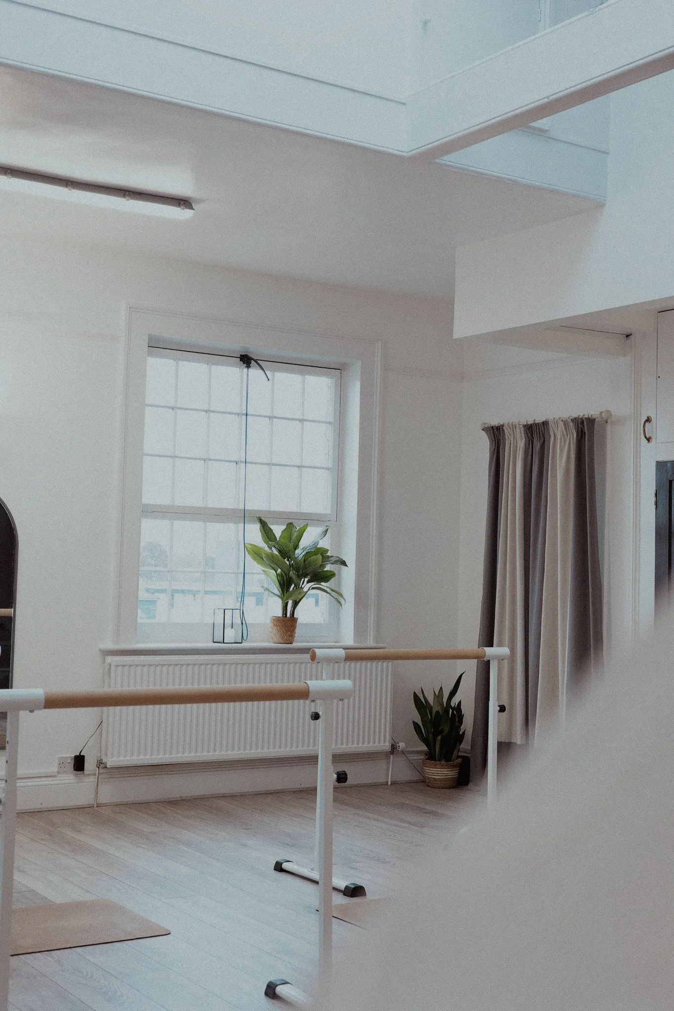 A room with white walls, light wood flooring, a window with curtains, a potted plant on the windowsill, and two ballet barres in the foreground.