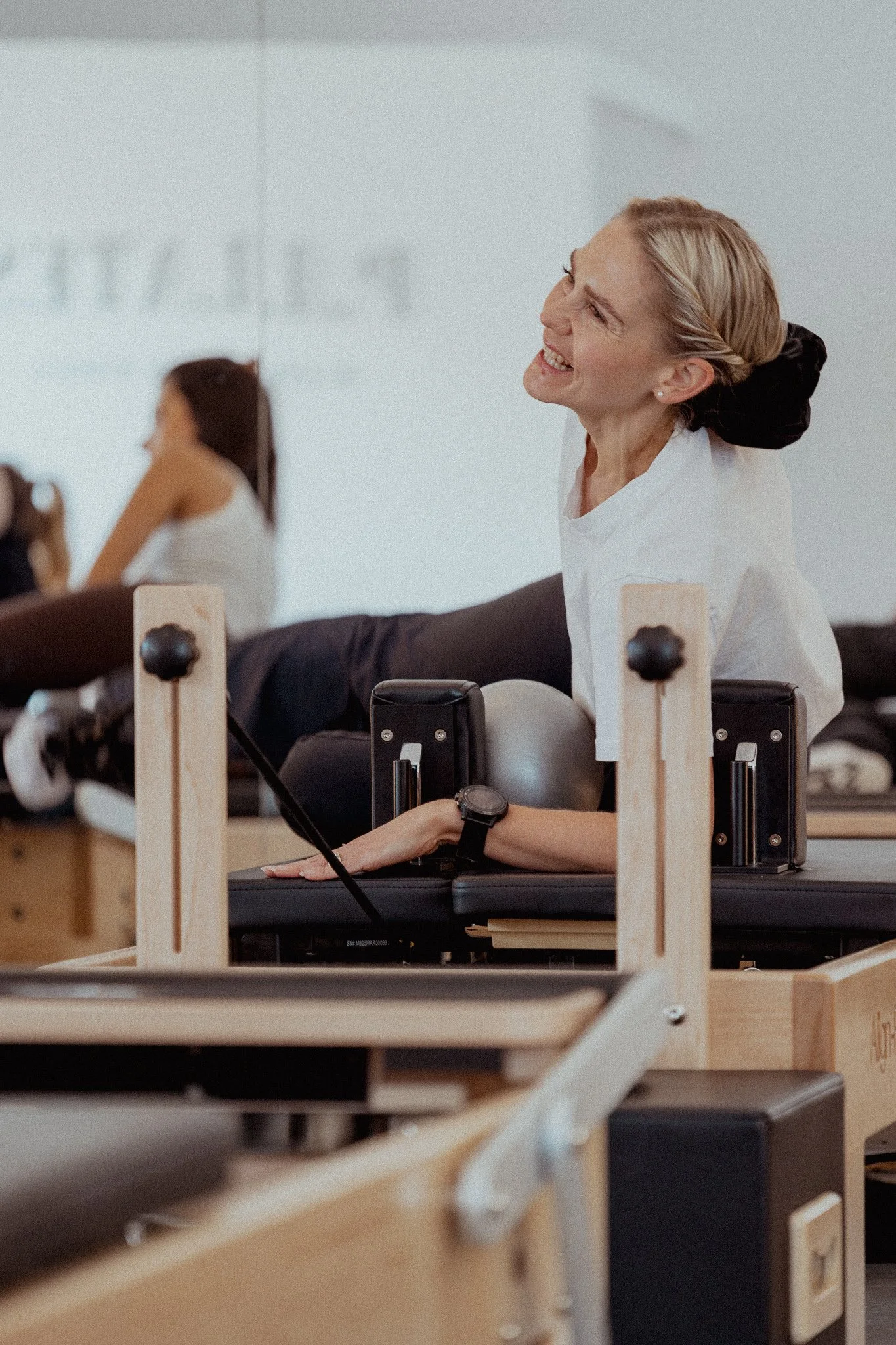 A woman with blonde hair, smiling, seated at a Pilates reformer machine in a fitness studio, with a glimpse of a woman in white shirt and black pants in the background.