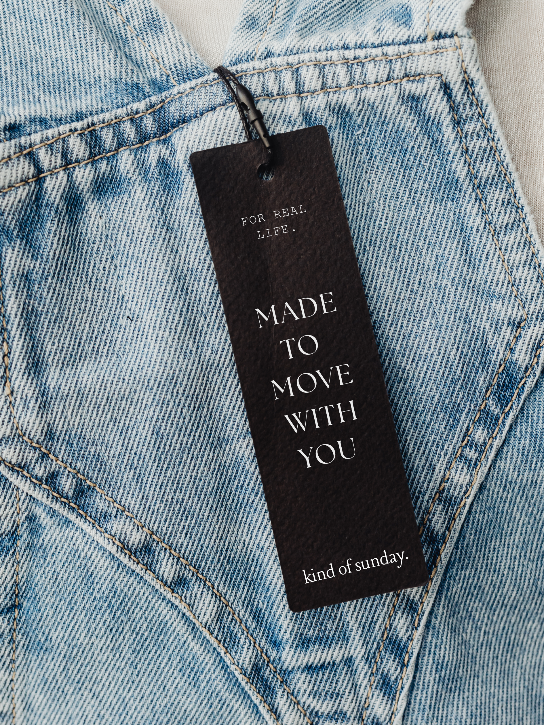 Close-up of a black clothing tag hanging from a pair of light blue denim jeans with orange stitching. The tag reads: 'FOR REAL LIFE. MADE TO MOVE WITH YOU. kind of sunday.'