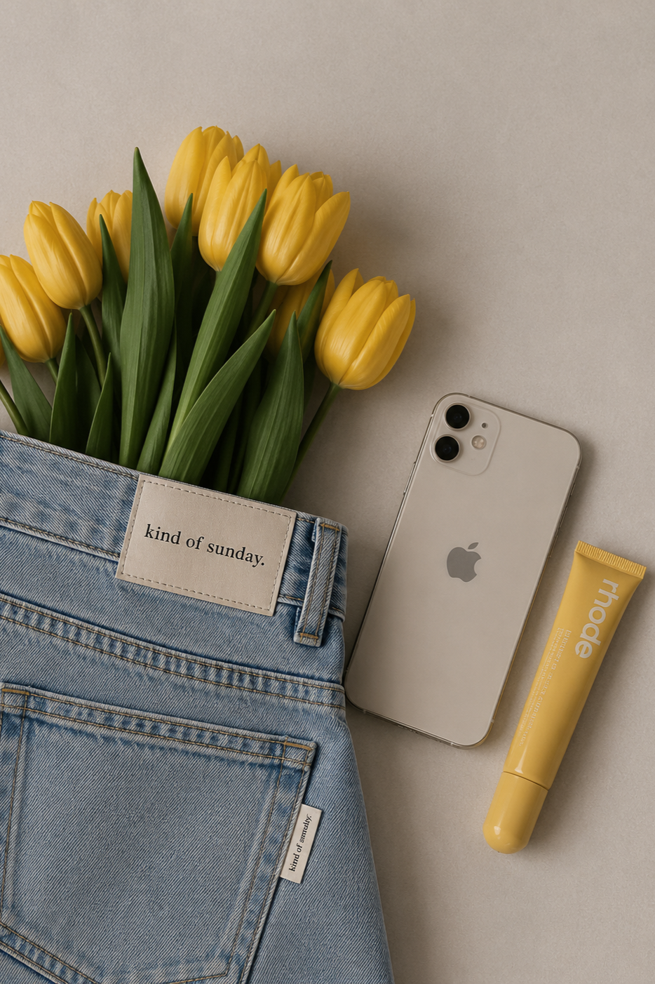 Yellow tulips in a denim pocket labeled 'kind of sunday,' an iPhone, and a tube of Rhode sunscreen on a light-colored surface.