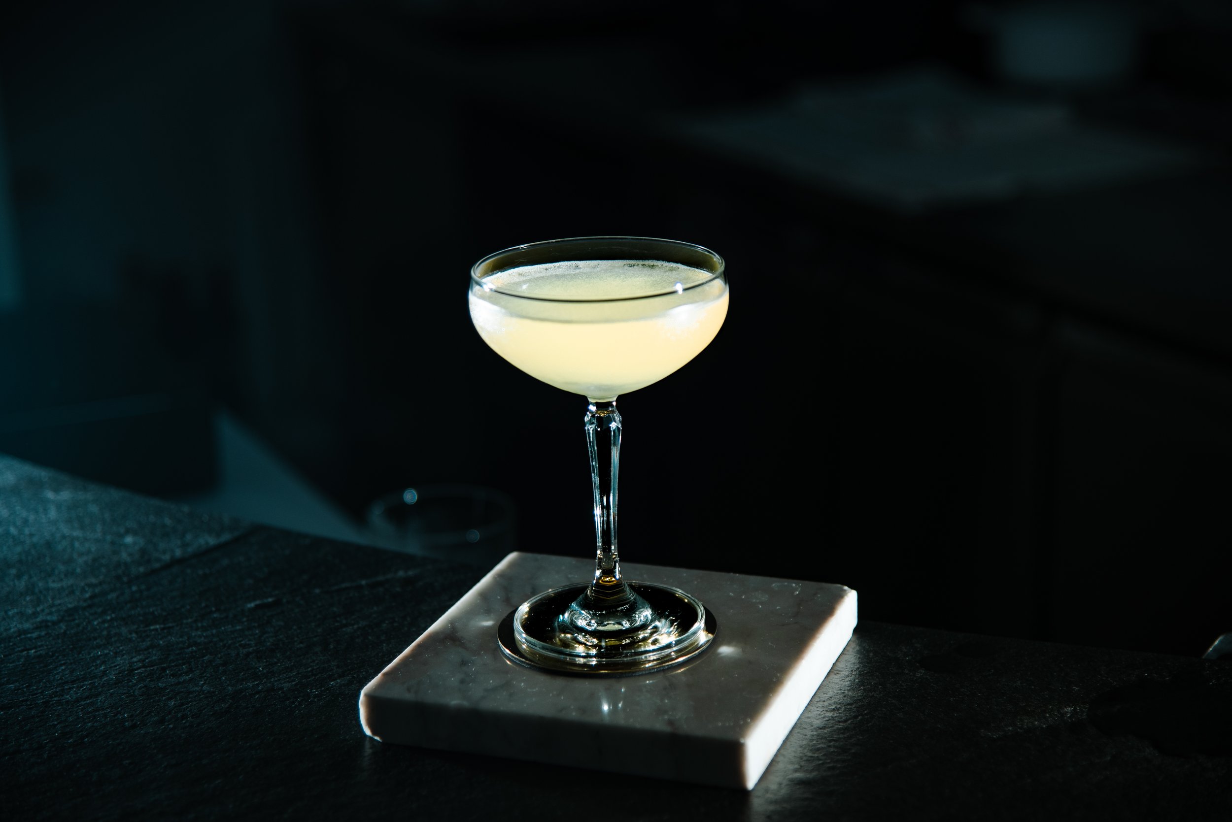A cocktail in a coupe glass with pale yellow liquid, on a white marble surface, against a dark background.