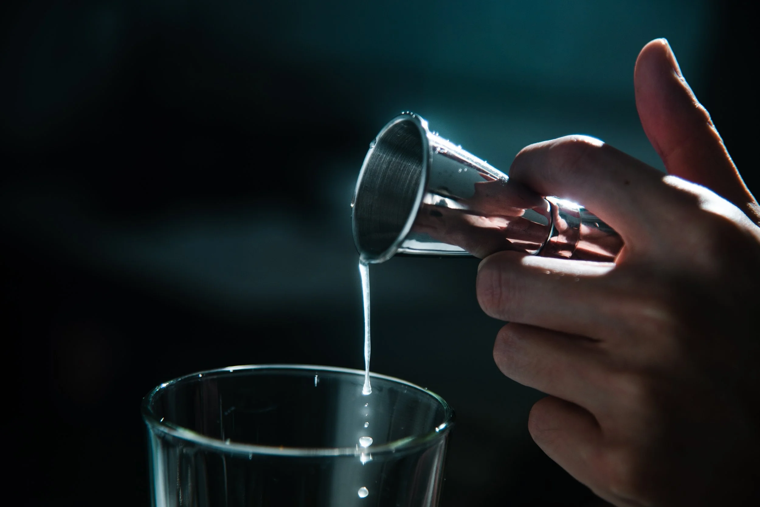 A hand holding a metal cup pouring liquid into a glass.