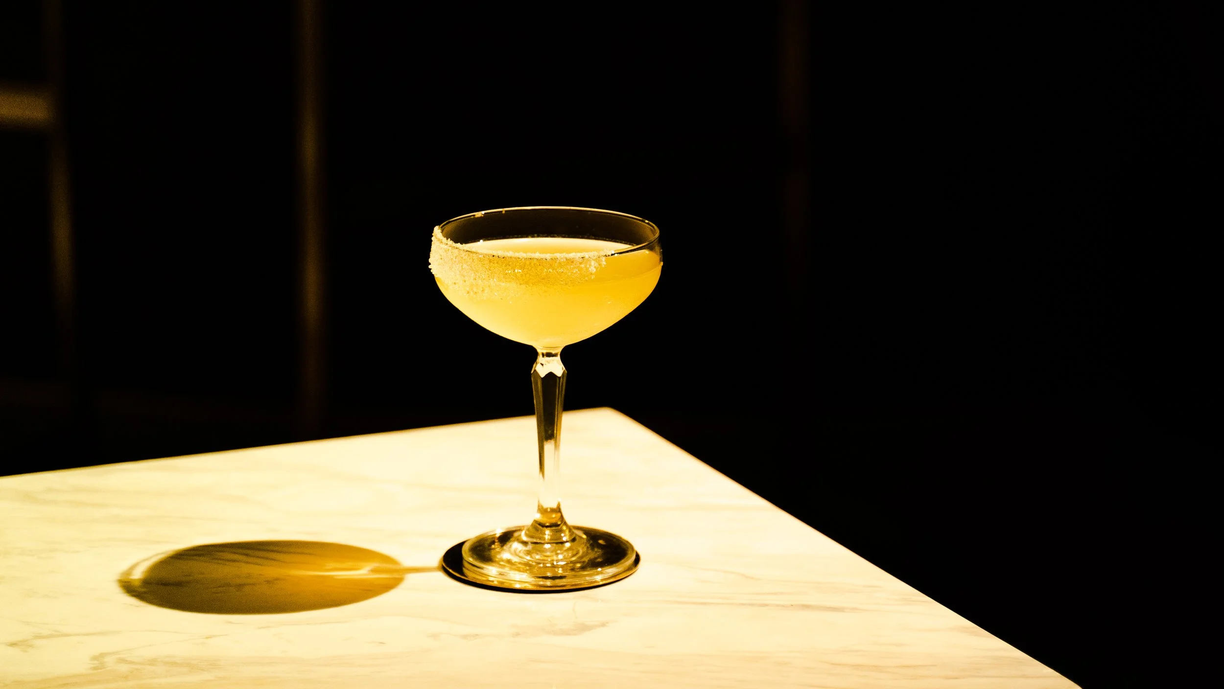 A yellow cocktail in a coupe glass with a sugared rim, placed on a wooden surface with a dark background.